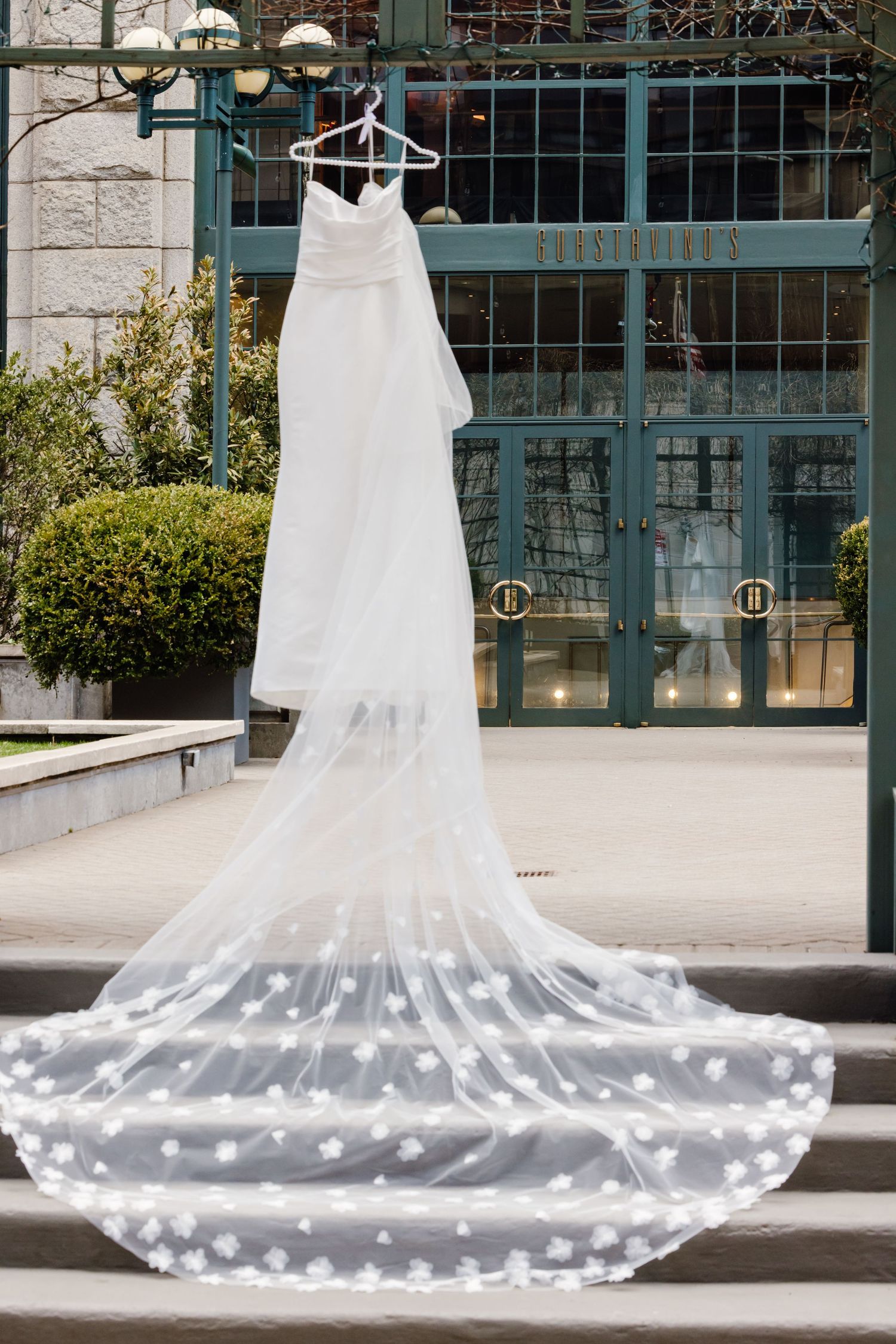 Strapless white wedding gown with floral veil train displayed on hanger outside building entrance.