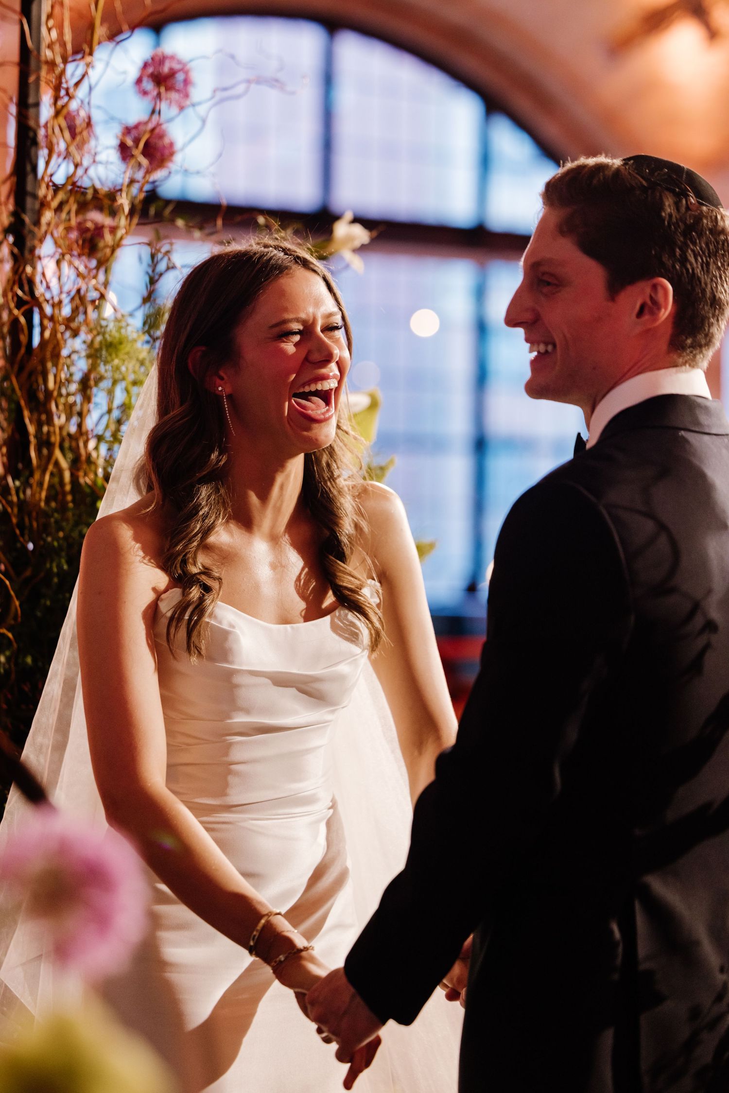 Laughing bride in white strapless gown holds hands with smiling groom at wedding ceremony altar.