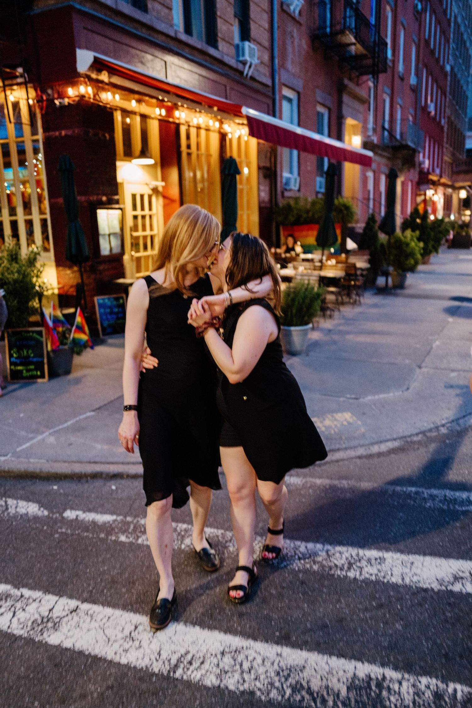 Two women in black dresses kissing on a charming NYC street at dusk.