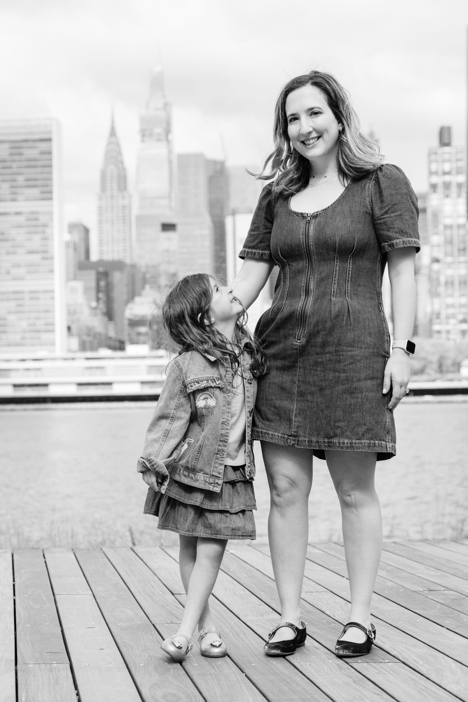Black and white photo of mother and daughter standing together on a boardwalk with NYC skyline.