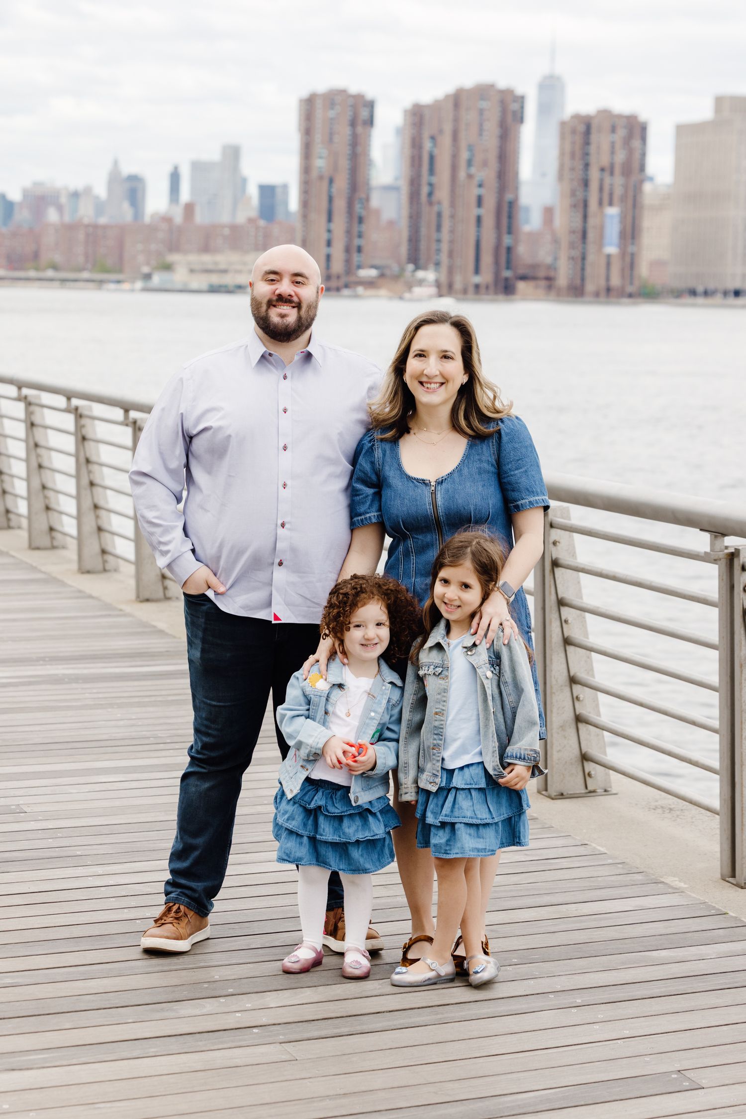 Family of four posing on a waterfront boardwalk with New York City skyline in the background.