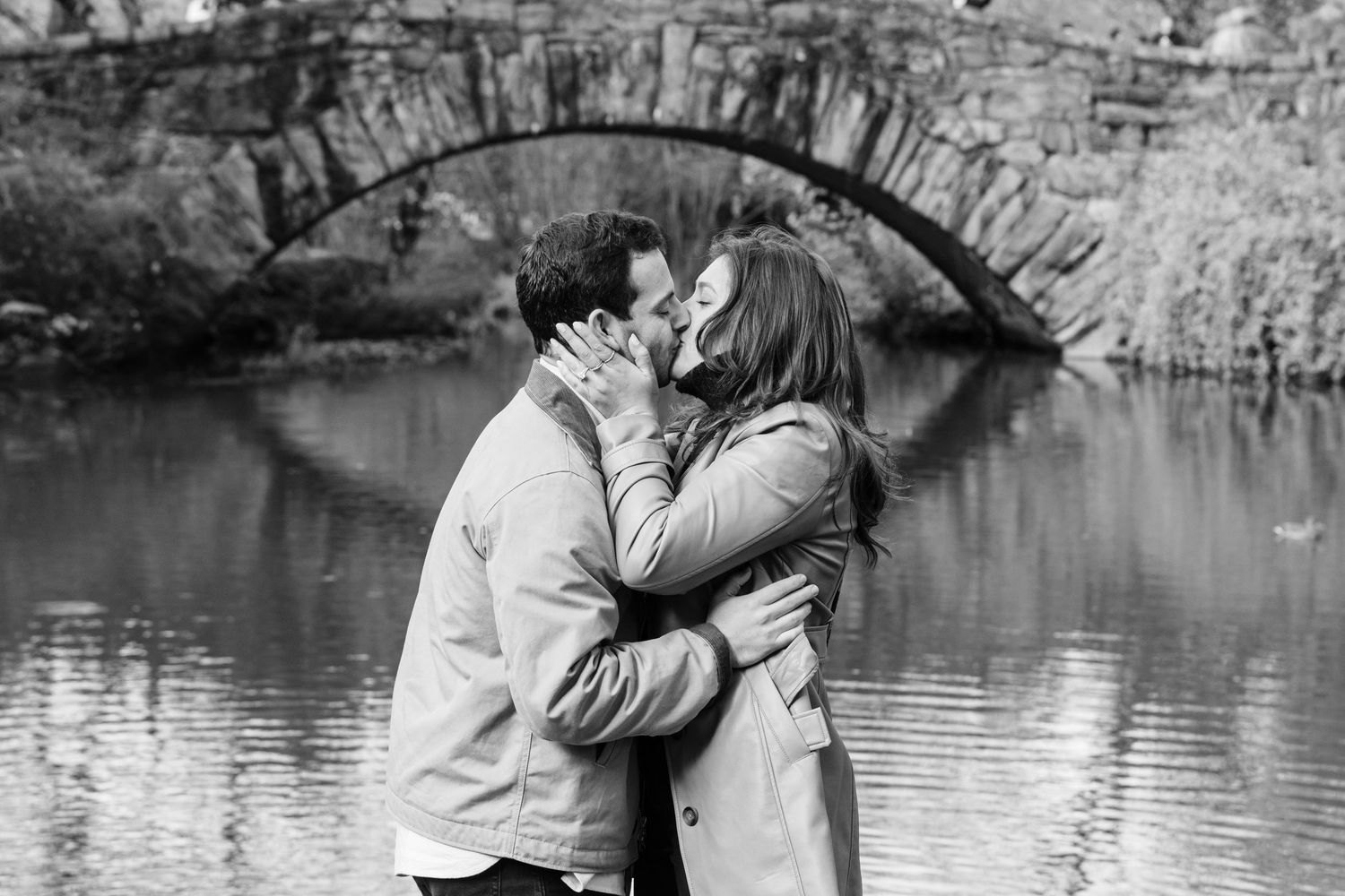 Black and white photo of a couple sharing a romantic kiss near a stone bridge over a serene pond.