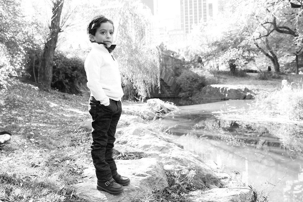 Black and white photo of a young child standing outdoors near a pond, wearing a white shirt and dark pants.