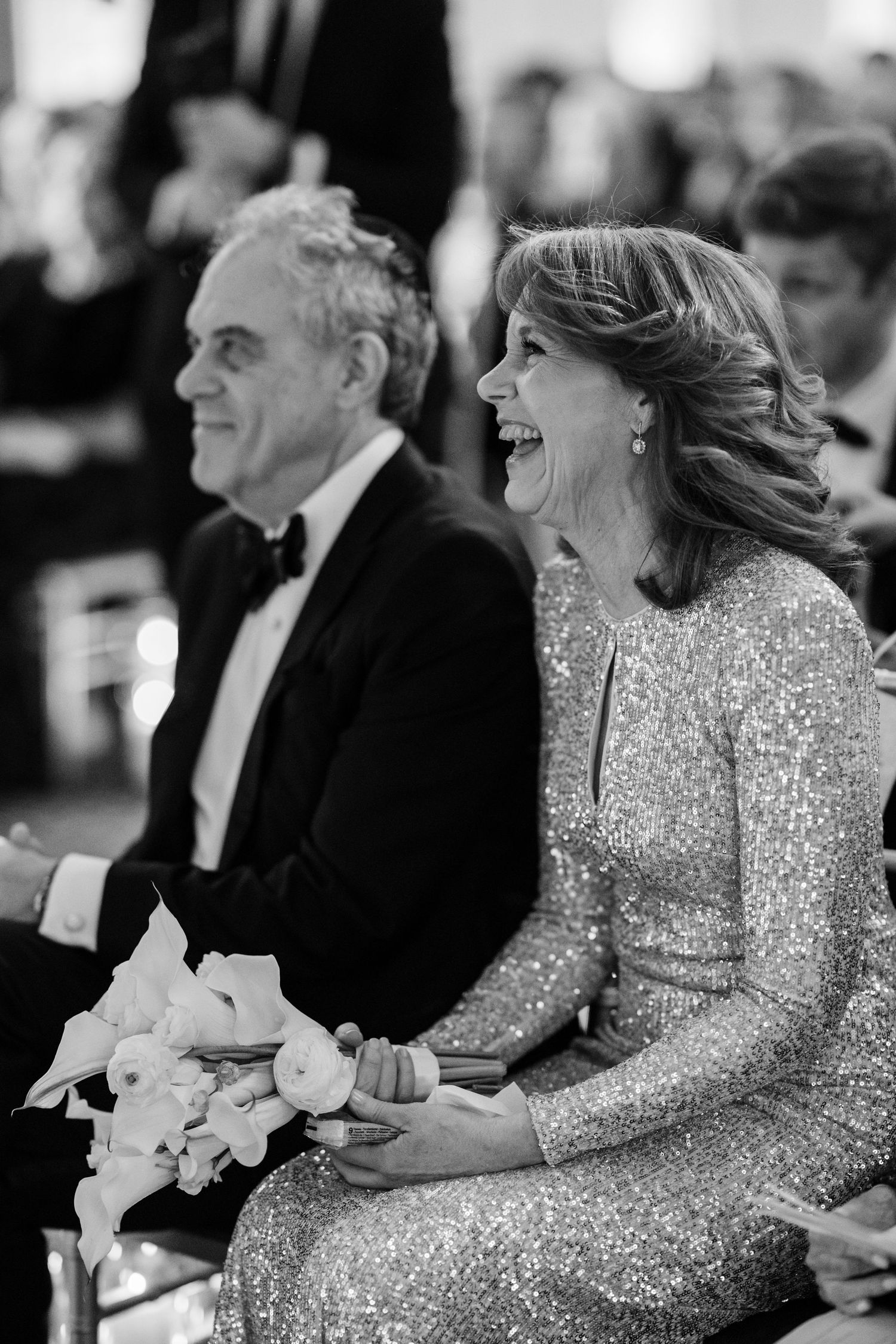 Joyful wedding guests in formal attire laugh during ceremony, woman in sequin gown holds bridal bouquet.