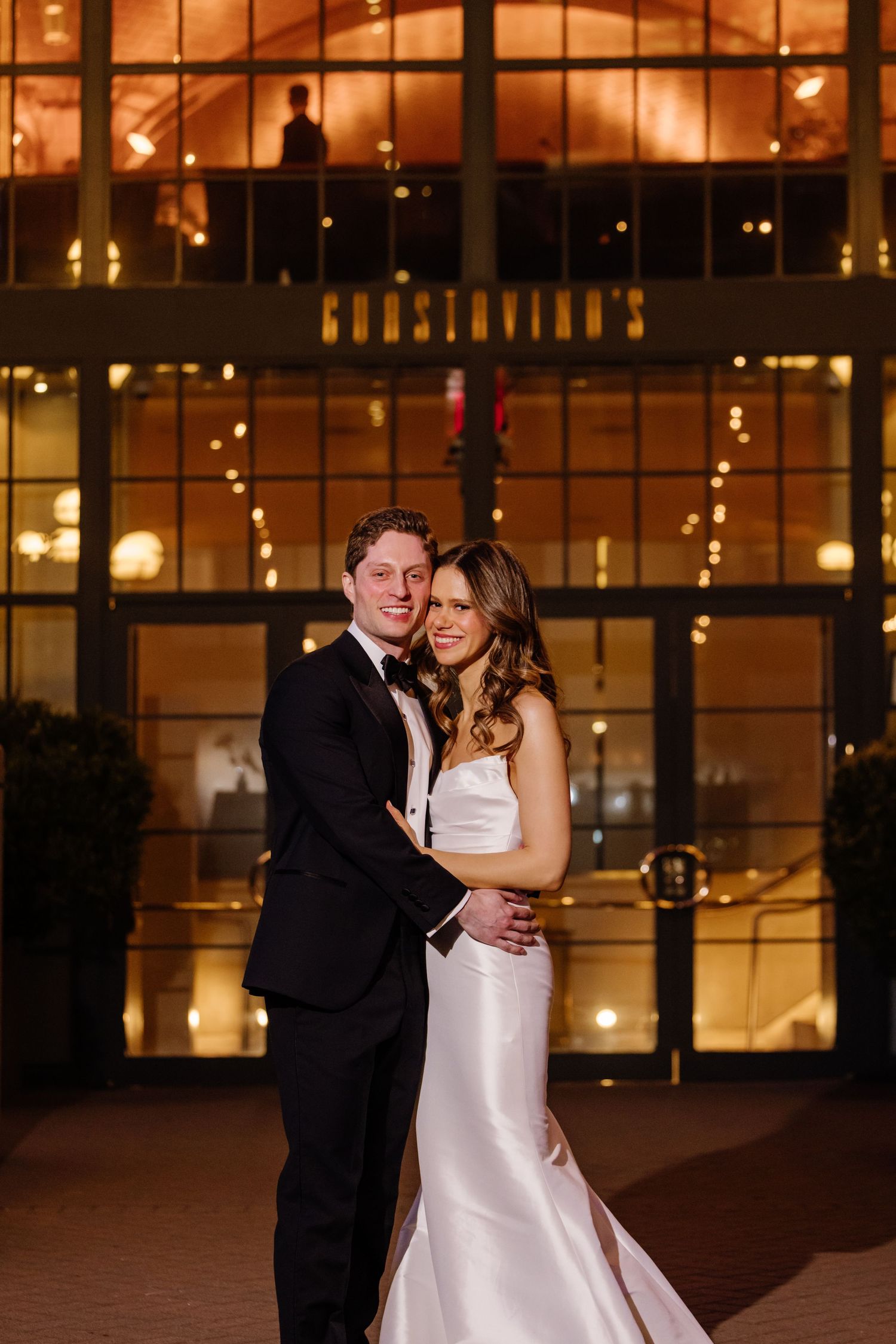 Bride and groom pose together smiling outside Guastavino's venue glowing warmly at night.