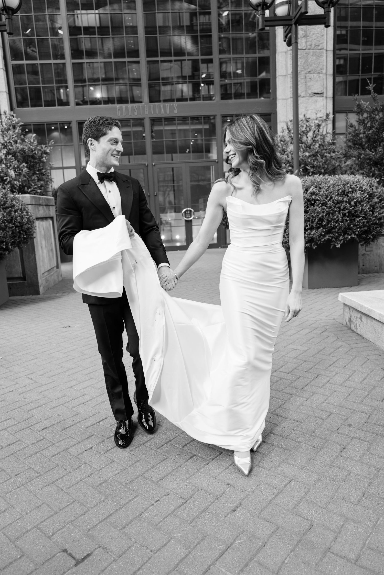 Black and white photo of smiling newlyweds holding hands and walking on cobblestone street outdoors.