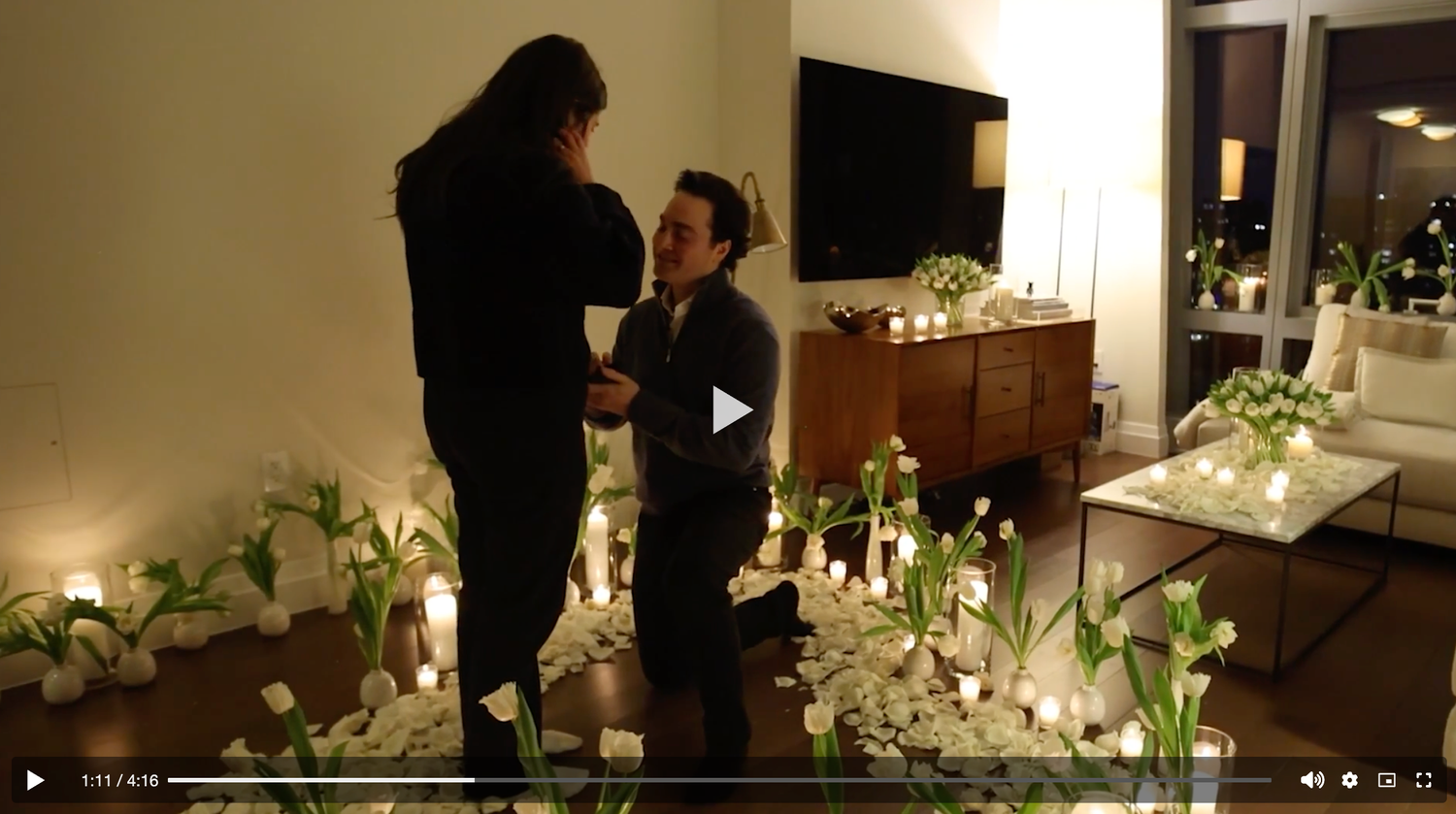 Romantic candlelit marriage proposal with white floral arrangements in a softly lit room.
