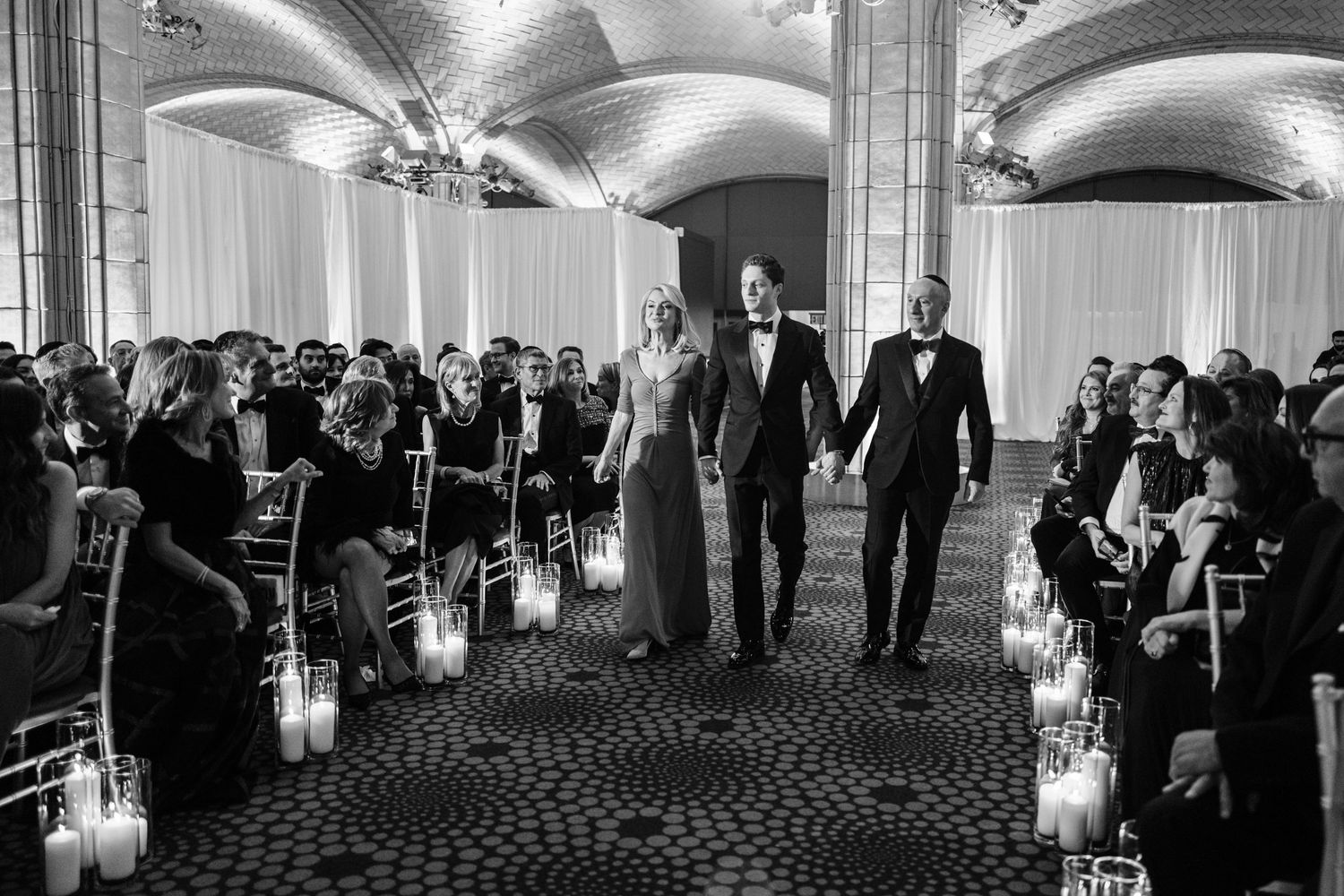Bride and groom walk down candlelit aisle at elegant wedding ceremony while guests look on.
