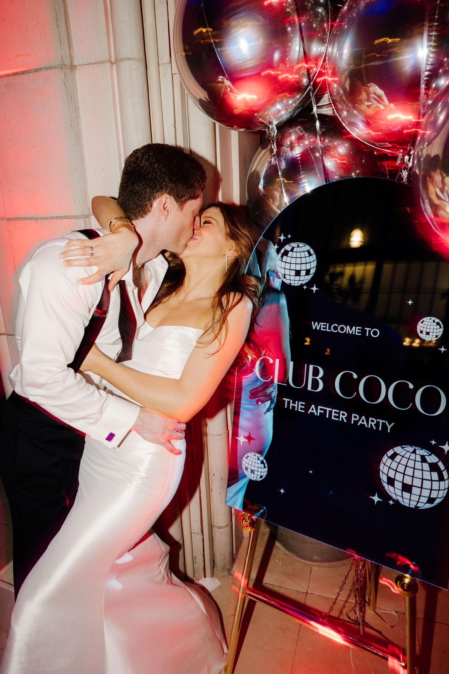 Newlyweds kissing beside a Club Coco after party sign with metallic balloons at their wedding.