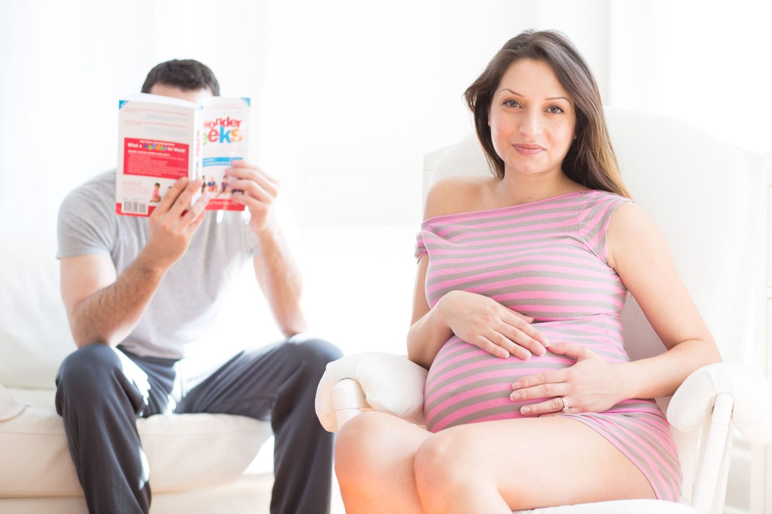 Pregnant woman in striped dress holds belly while man reads baby book on white sofa at home.