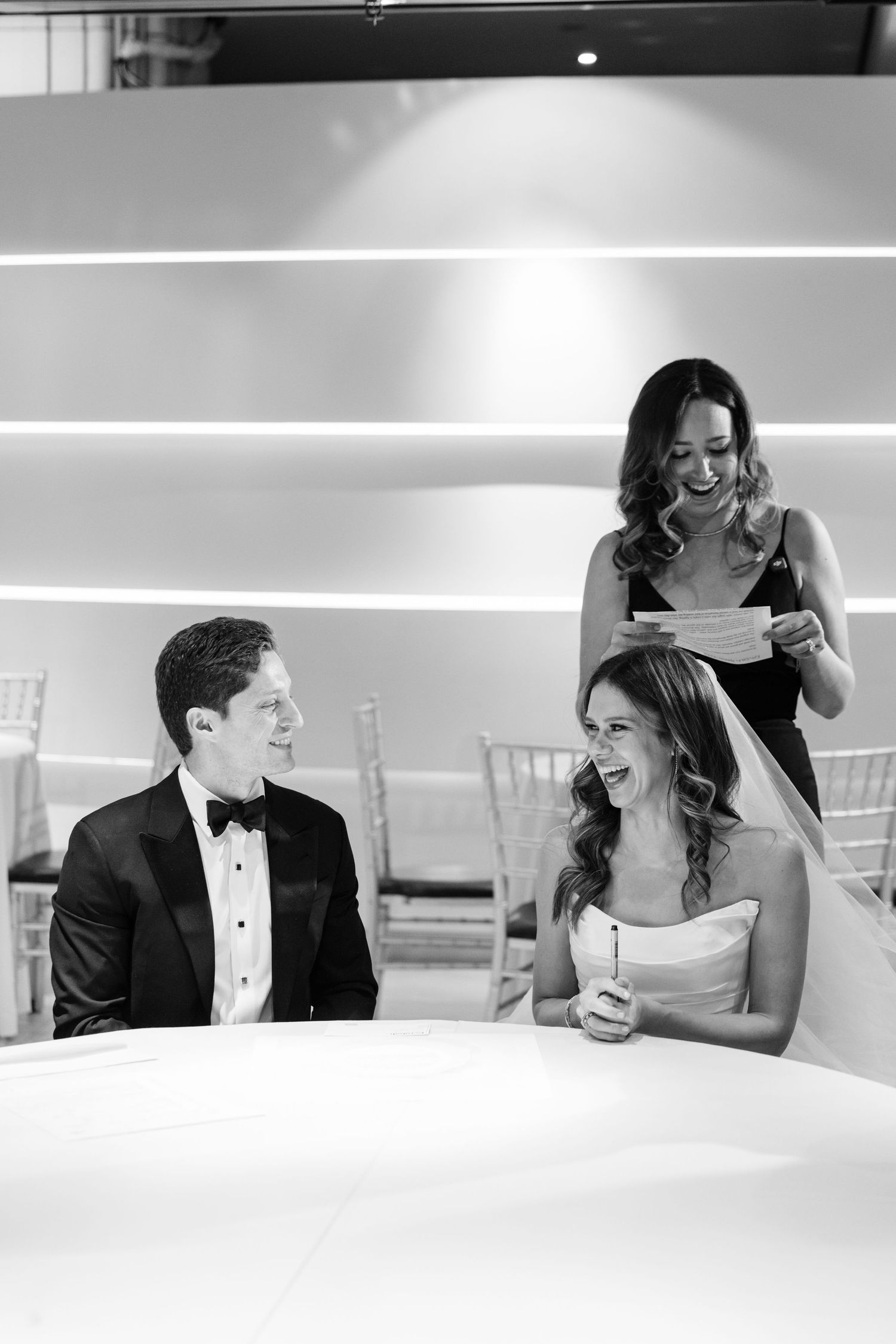 Black and white photo of bride and groom laughing during wedding speech at reception table.