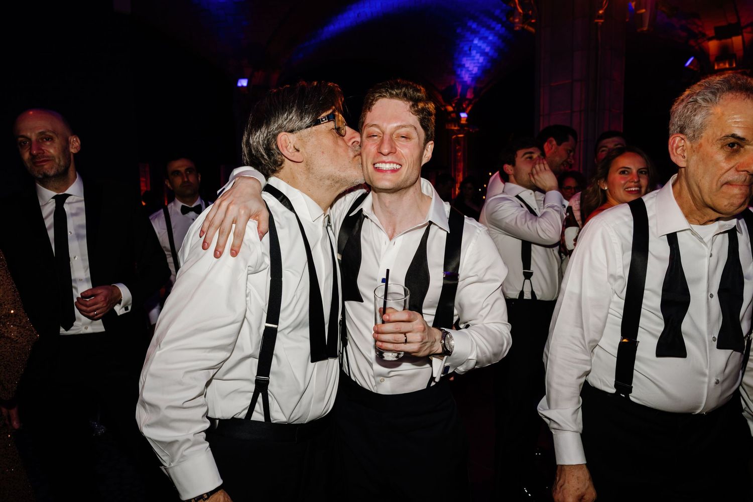 Two men in white shirts and suspenders sharing a celebratory kiss at a wedding reception.