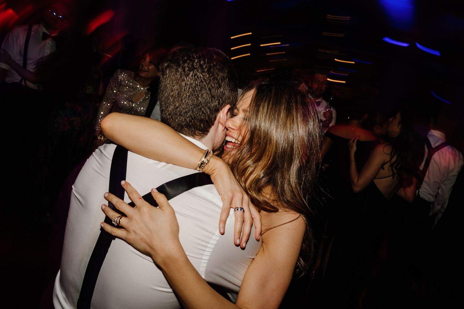 Couple sharing a passionate kiss on a crowded wedding reception dance floor with colorful lights.