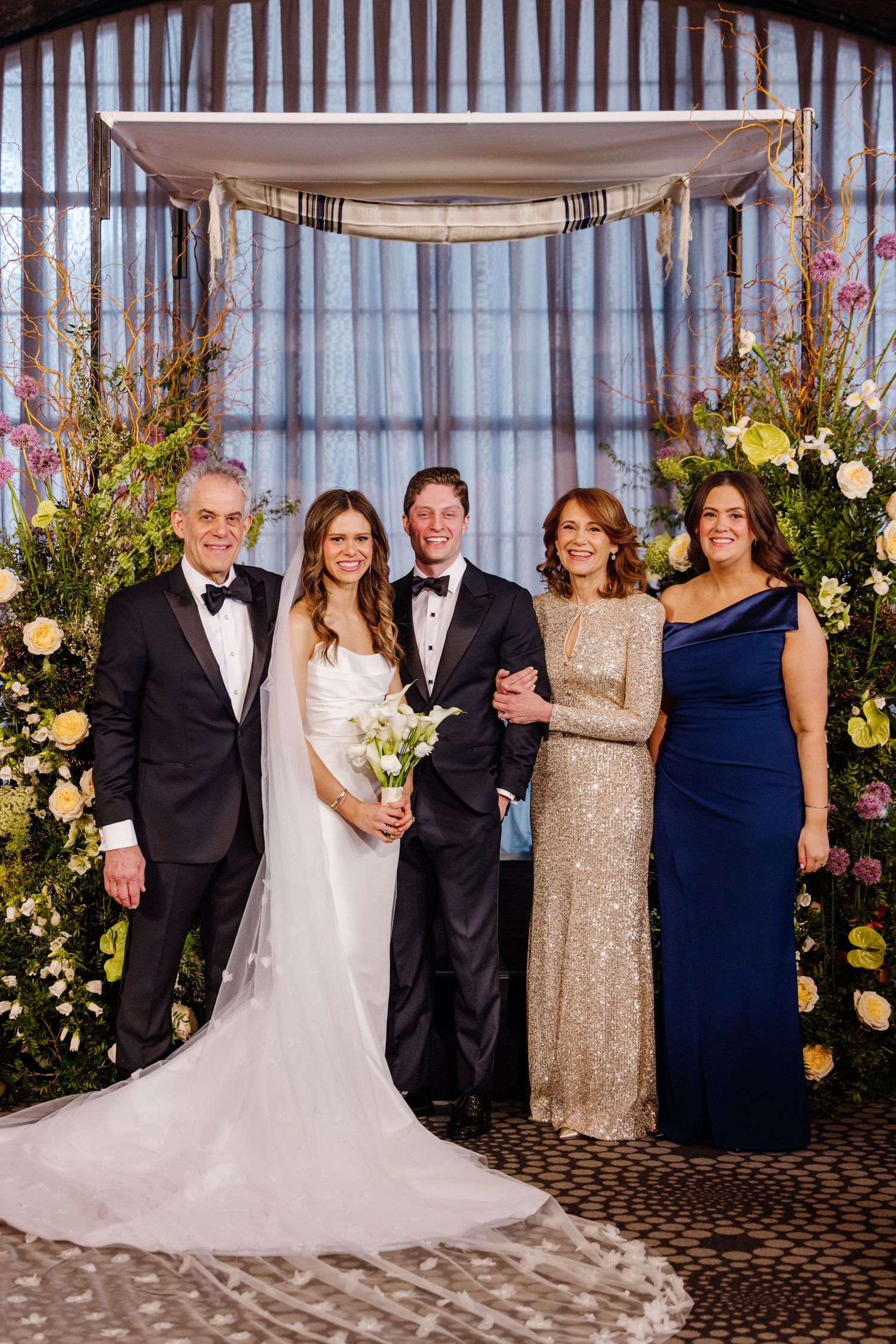 Bride and groom with four family members posing under floral chuppah at wedding ceremony.