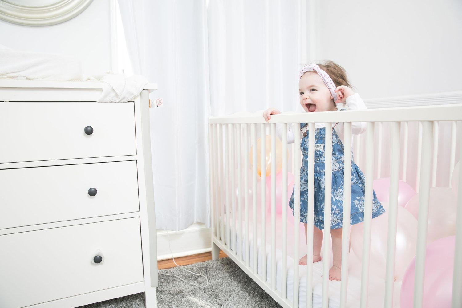 Smiling toddler girl in blue dress standing in white crib next to white dresser in bright nursery room.