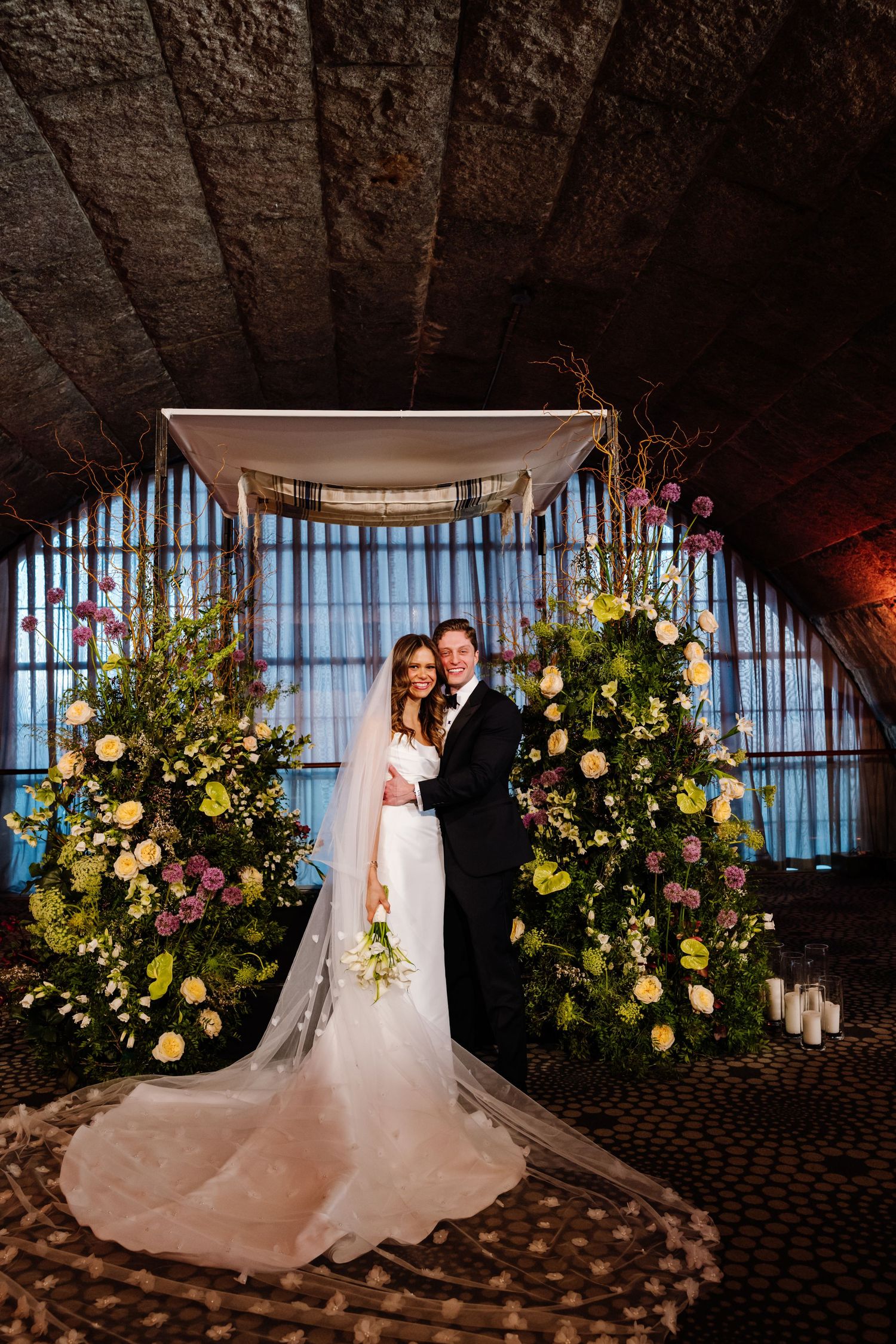 Bride and groom smiling under a floral chuppah adorned with colorful flowers in a stone venue.