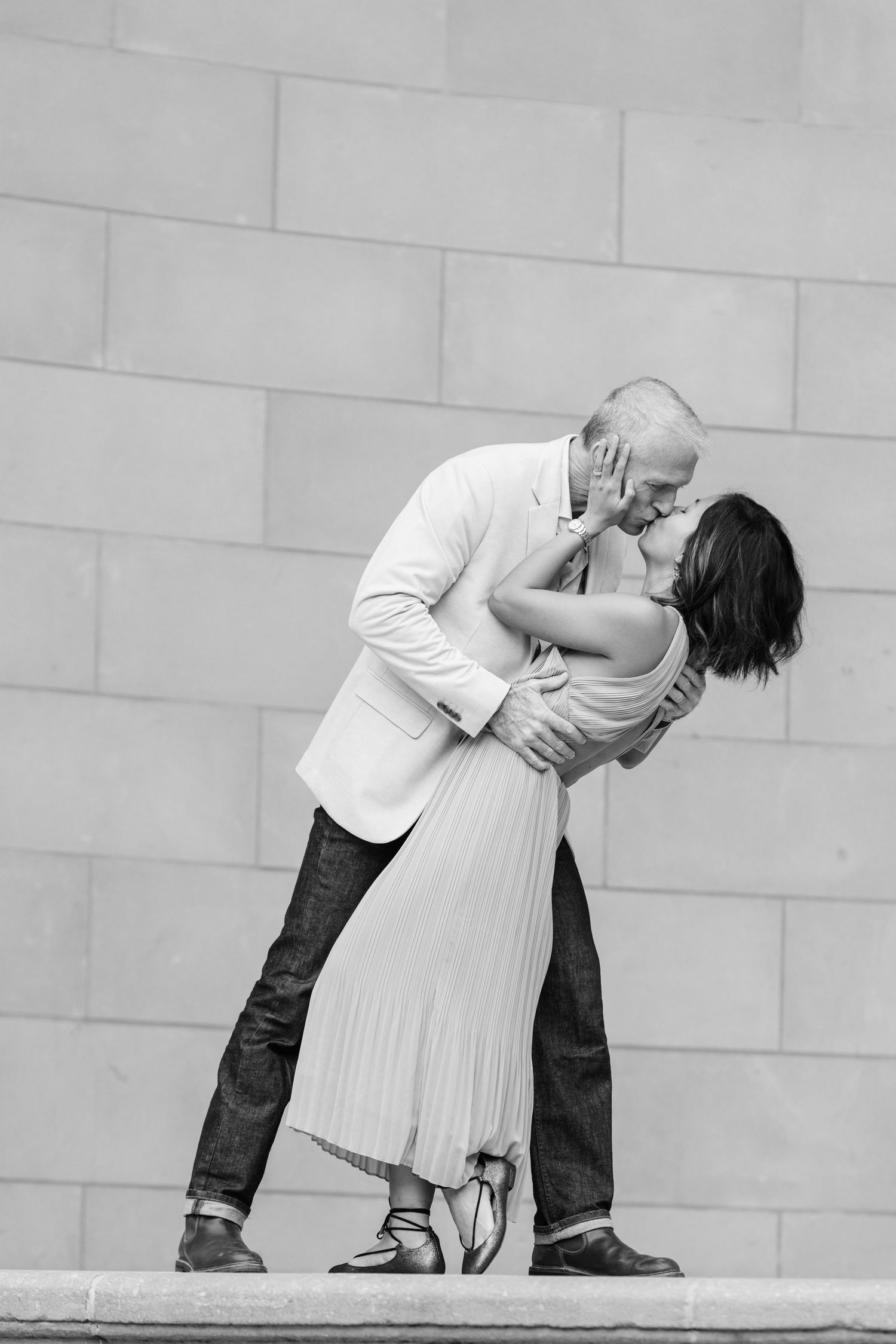 Black and white photo of a couple sharing a passionate dip kiss against a stone wall backdrop.