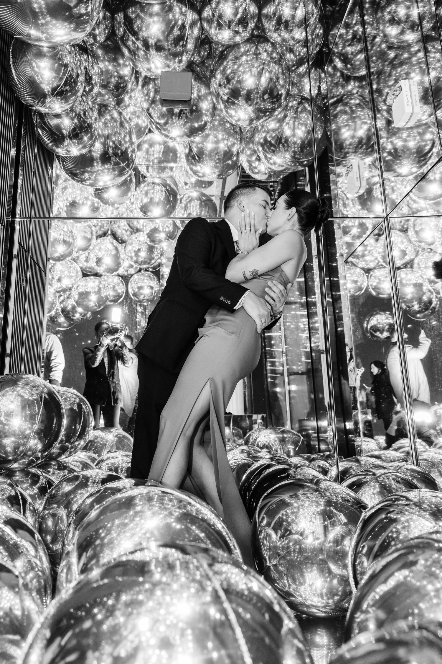 Black and white photo of couple kissing surrounded by shiny metallic balloons at party.