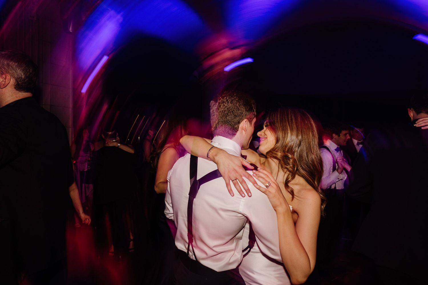 Couple sharing tender embrace on vibrant purple-lit wedding reception dance floor.