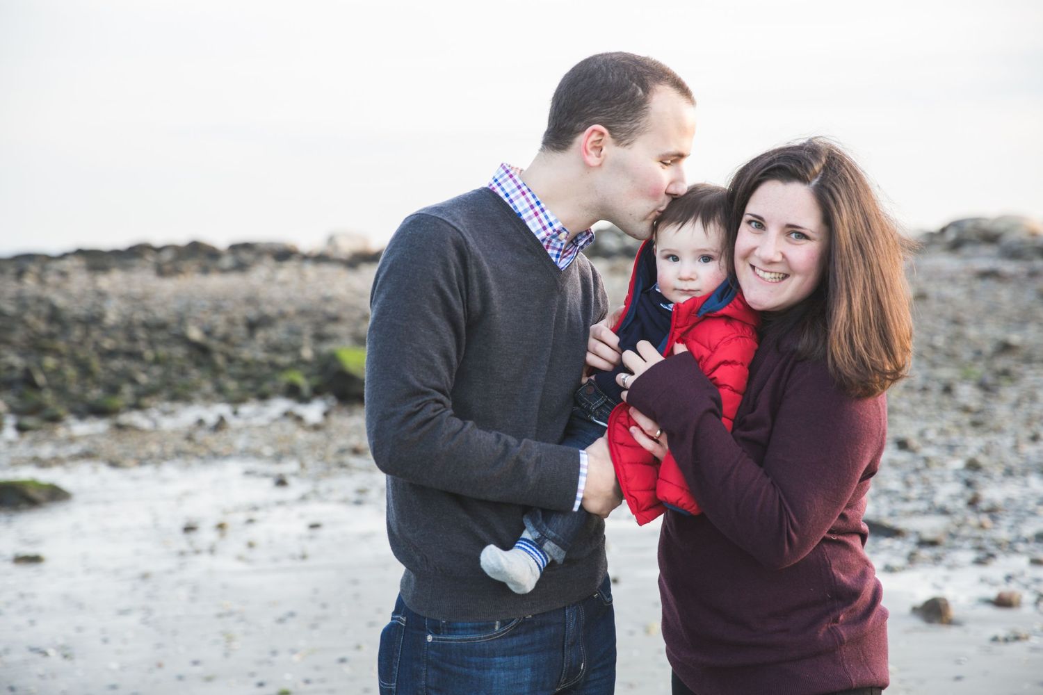 Happy family with baby posing on a rocky beach, parents smiling outdoors in casual fall attire.