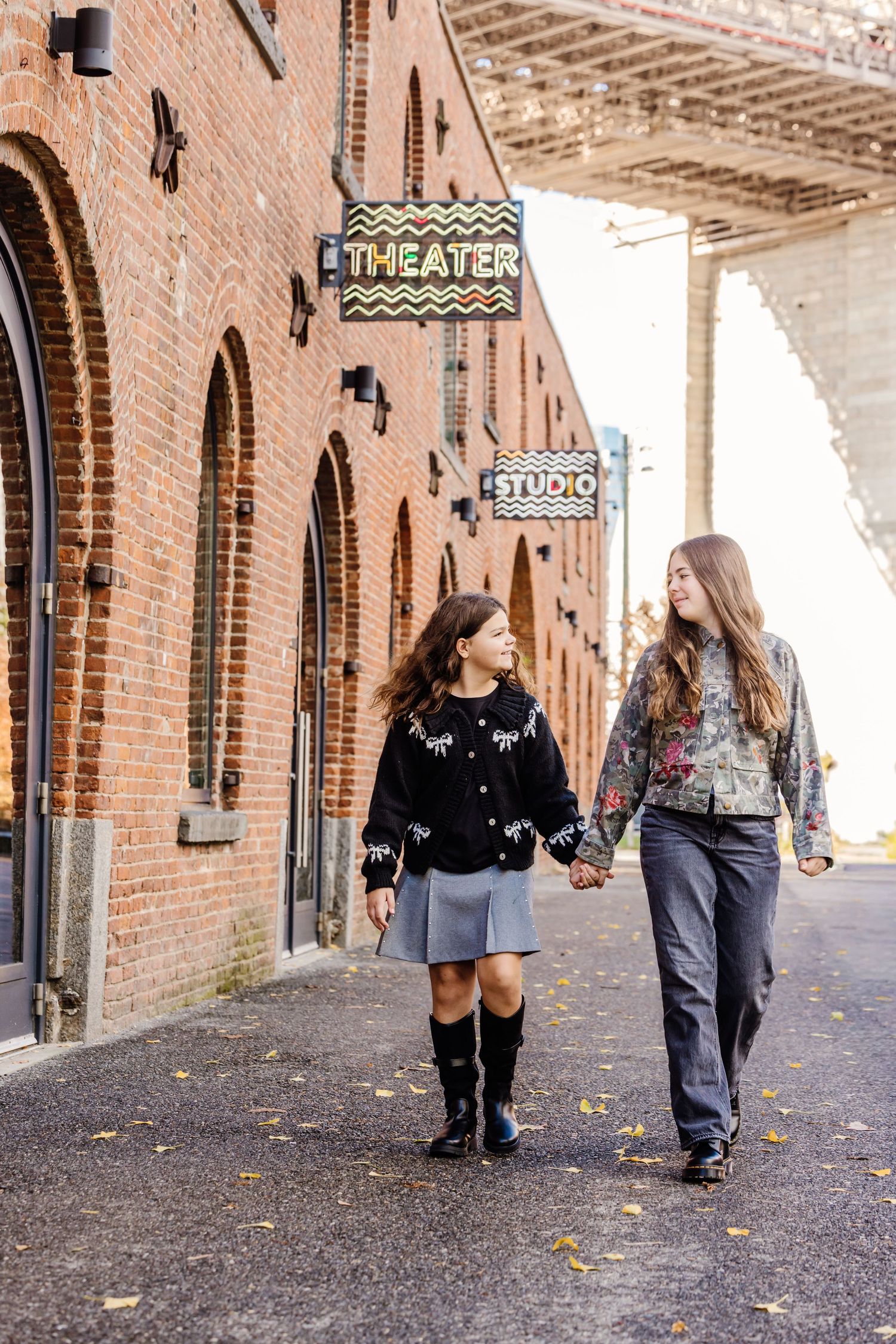 Two girls hold hands walking past a brick theater building with colorful signs in DUMBO Brooklyn.