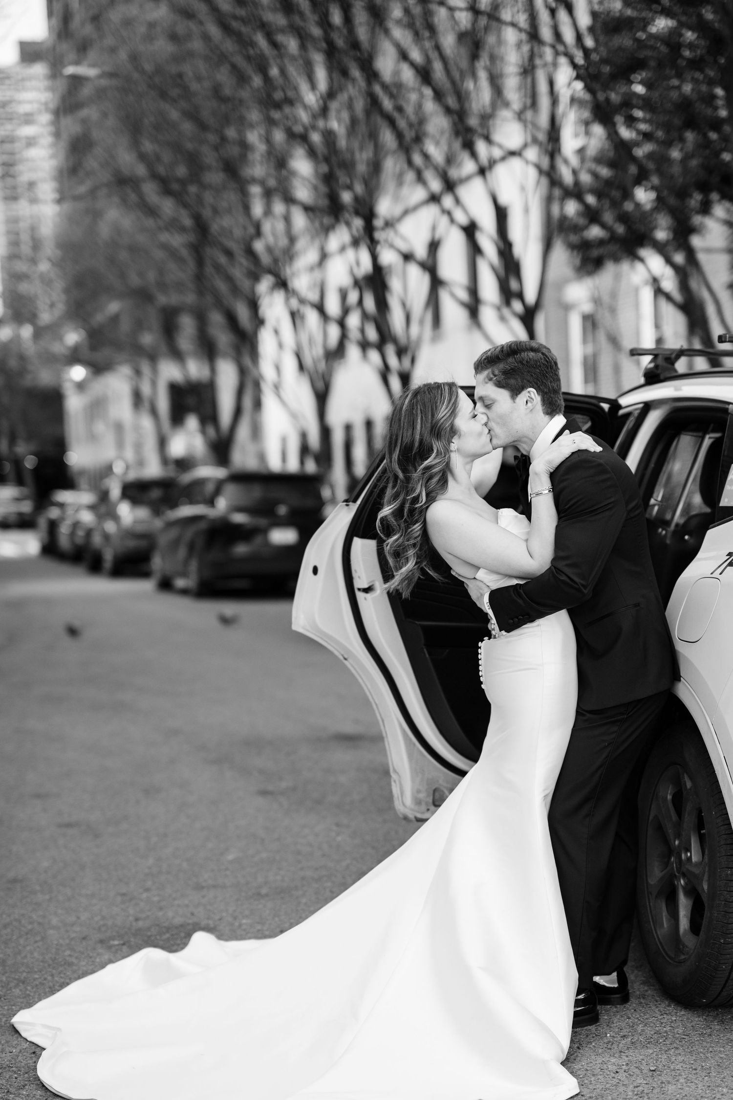 Black and white photo of a bride and groom sharing a kiss beside a car on a city street.