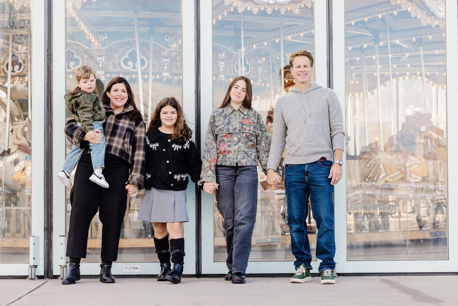 Family of five holding hands and smiling in front of a carousel at an outdoor venue.