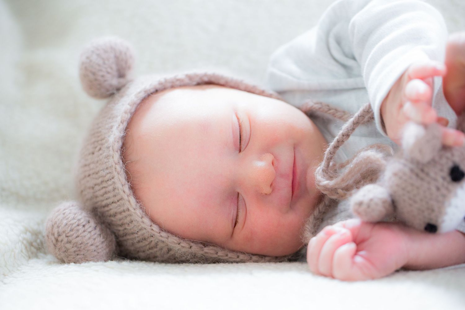 Sleeping newborn baby wearing a knit bear hat cuddles a stuffed toy on a soft white blanket.