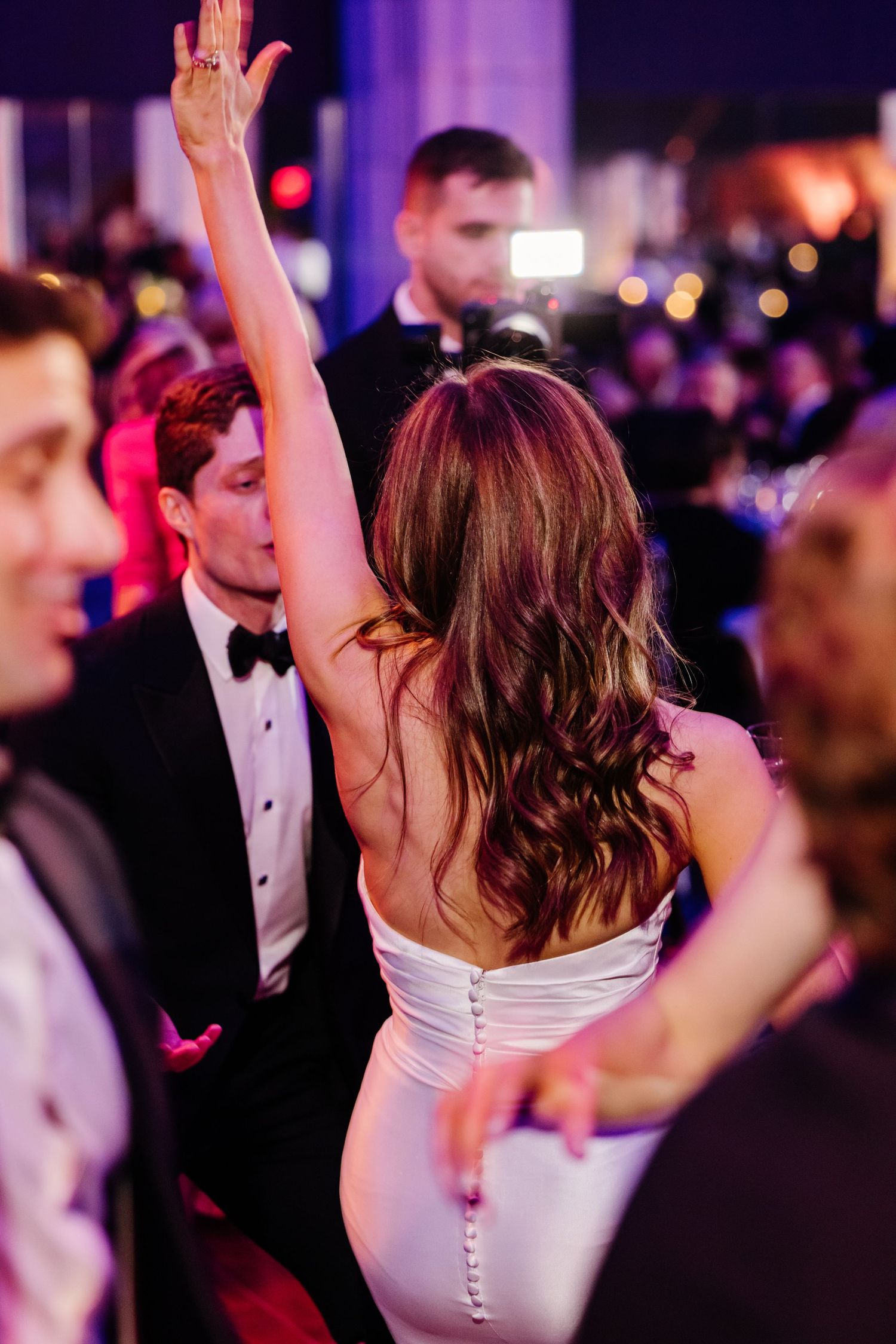 Bride in backless white gown dancing with arm raised at glamorous wedding reception.