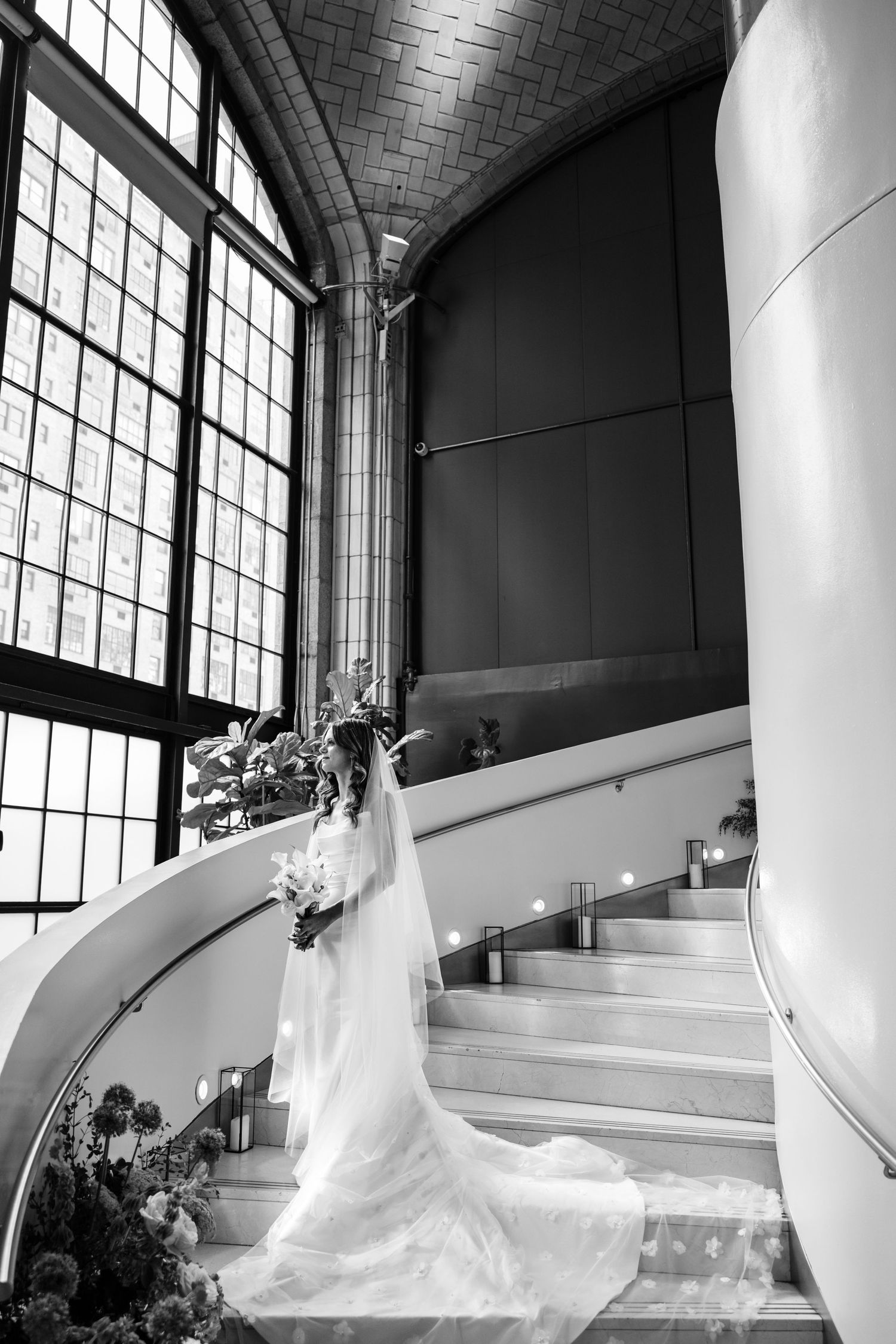 Black and white bridal portrait of bride in gown and veil on curved staircase by arched windows.