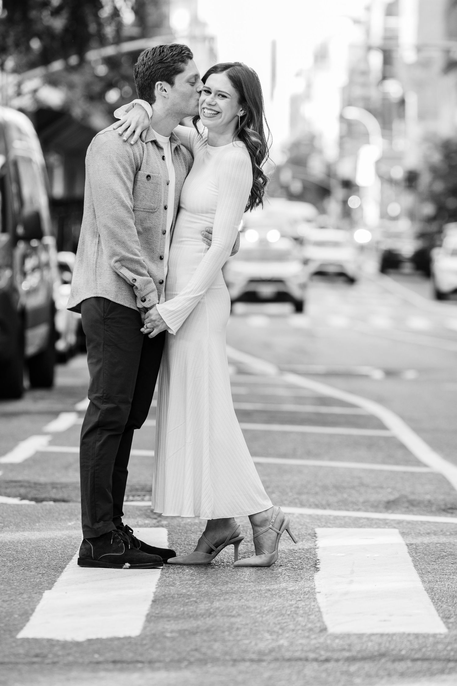 Black and white photo of a couple embracing on a New York City crosswalk in urban street setting.