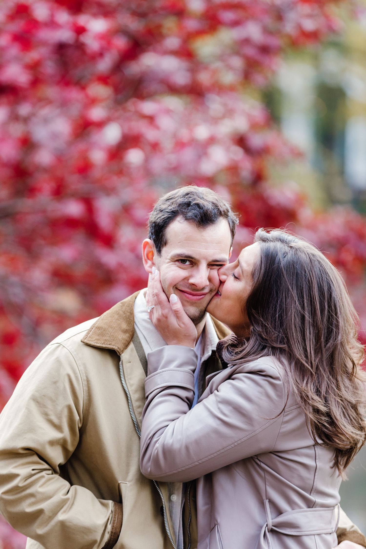 Woman kissing man on the cheek while he smiles in front of vibrant red autumn foliage.