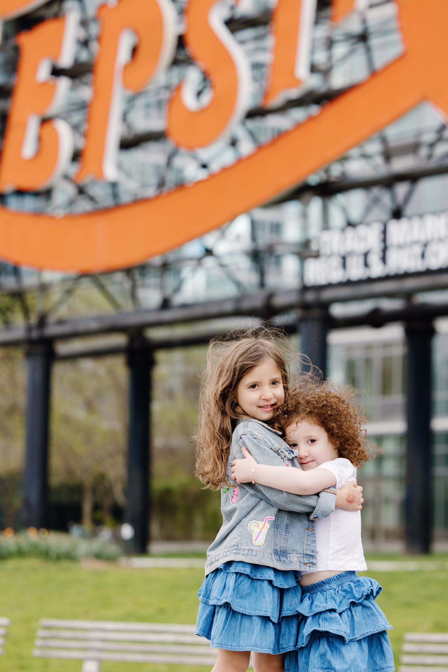 Two young sisters in matching denim outfits hugging in front of a colorful Pepsi sign outdoors.