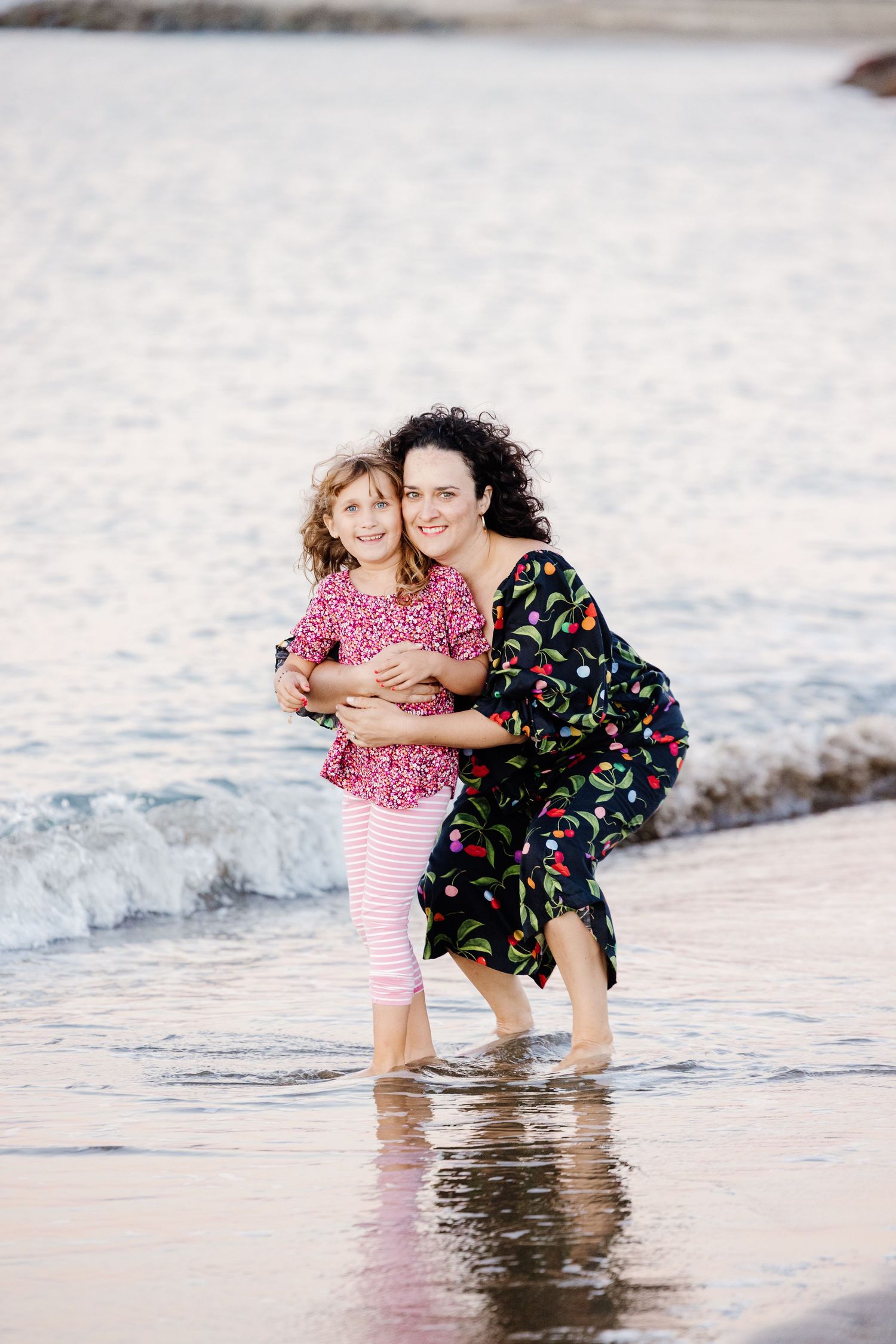 Mother with curly hair hugging her smiling young daughter while standing in shallow ocean waves at the beach.