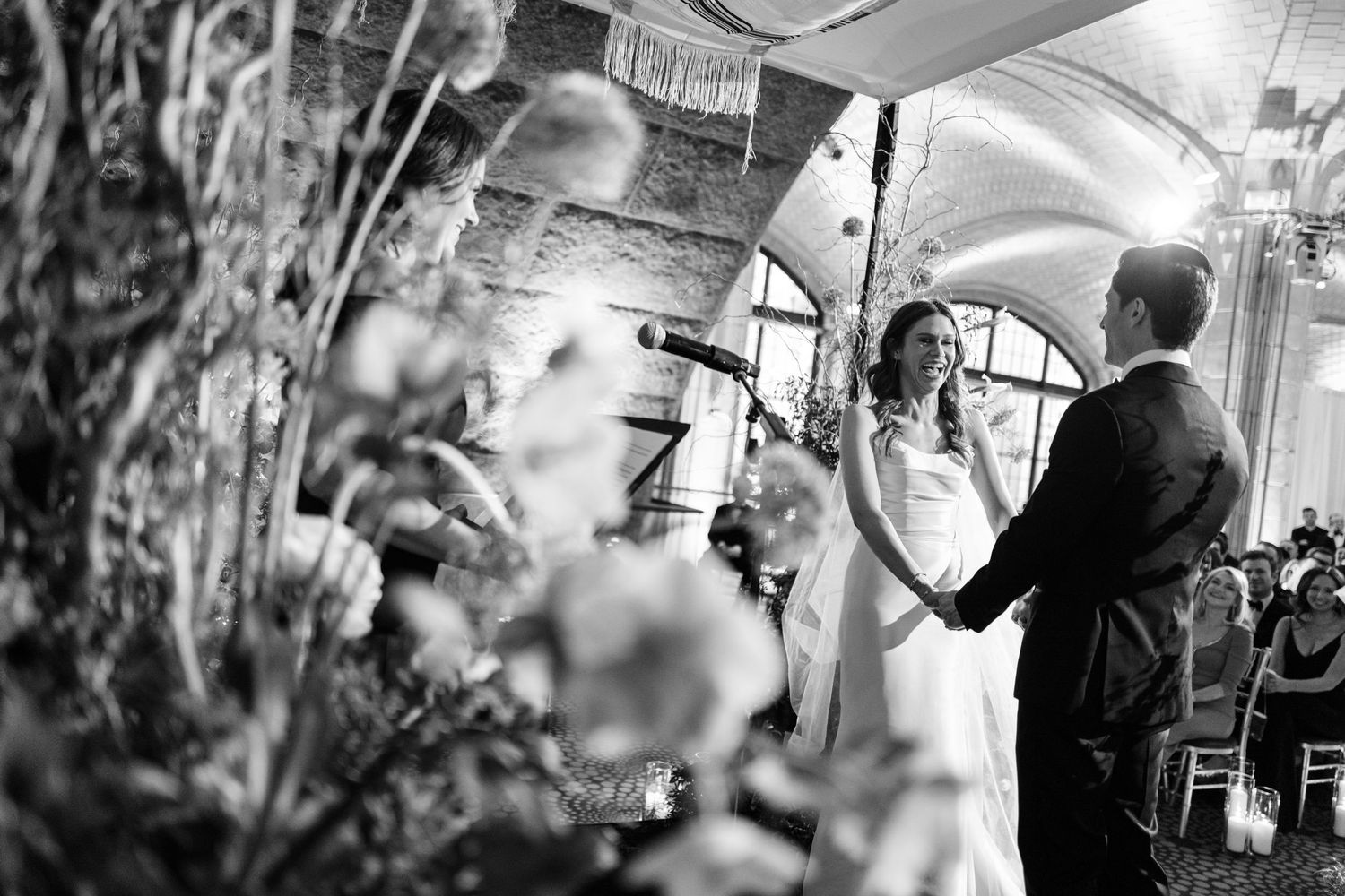 Bride and groom exchange vows at floral-decorated altar during intimate wedding ceremony, black and white.