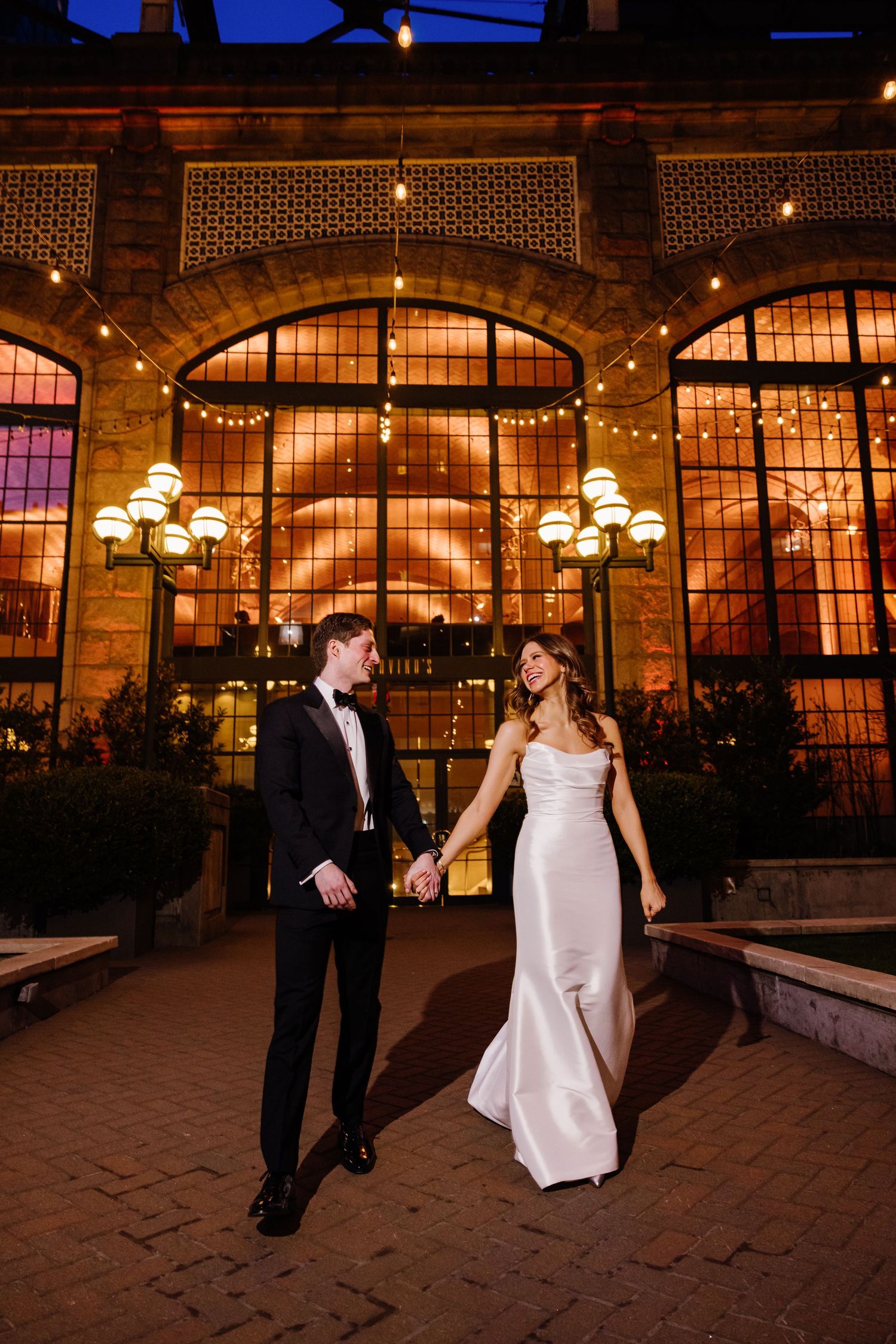 Bride and groom walk hand-in-hand outside illuminated Guastavino's venue at twilight with string lights.