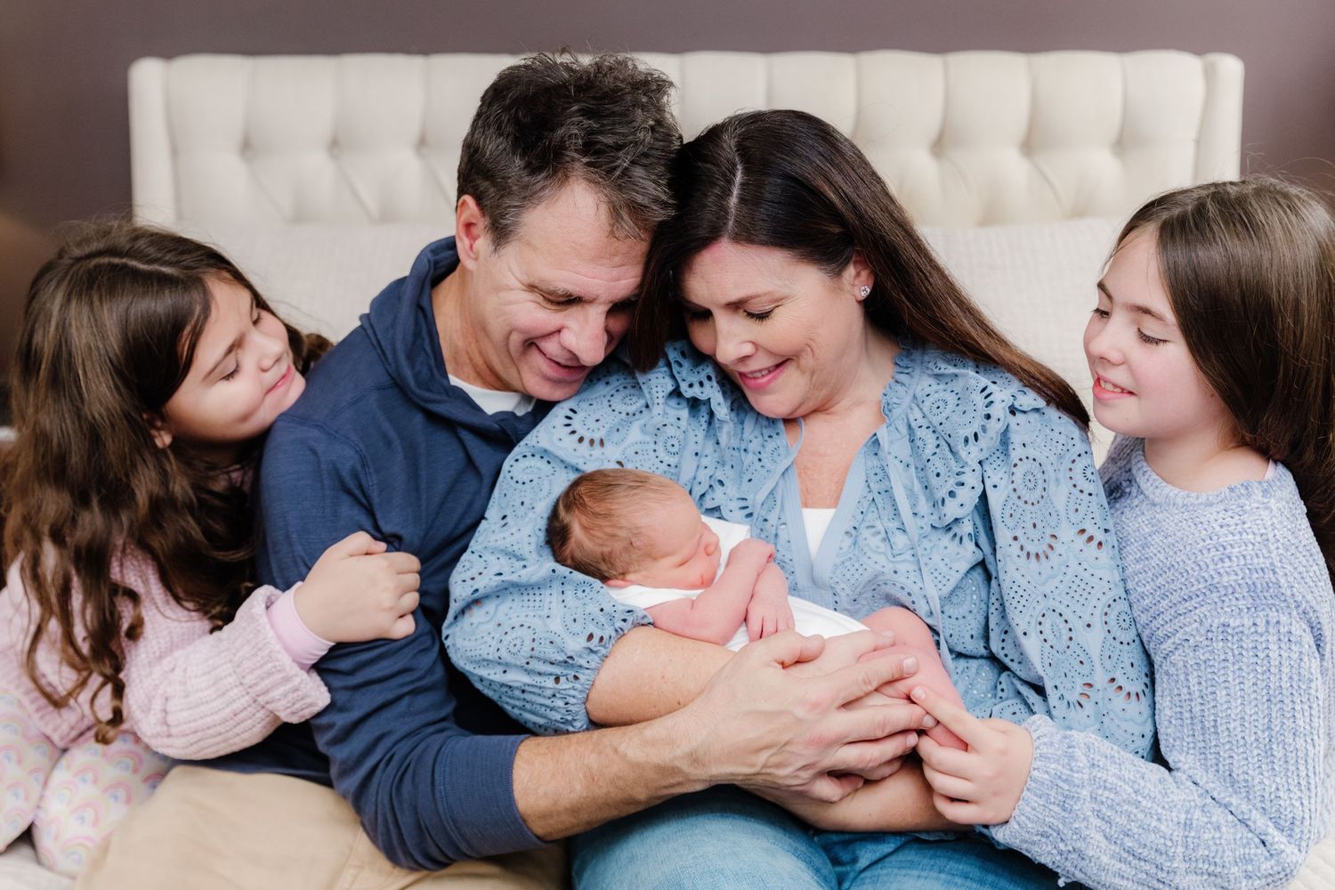 Happy family of five admiring their newborn baby together on a bed, with two young girls and smiling parents.