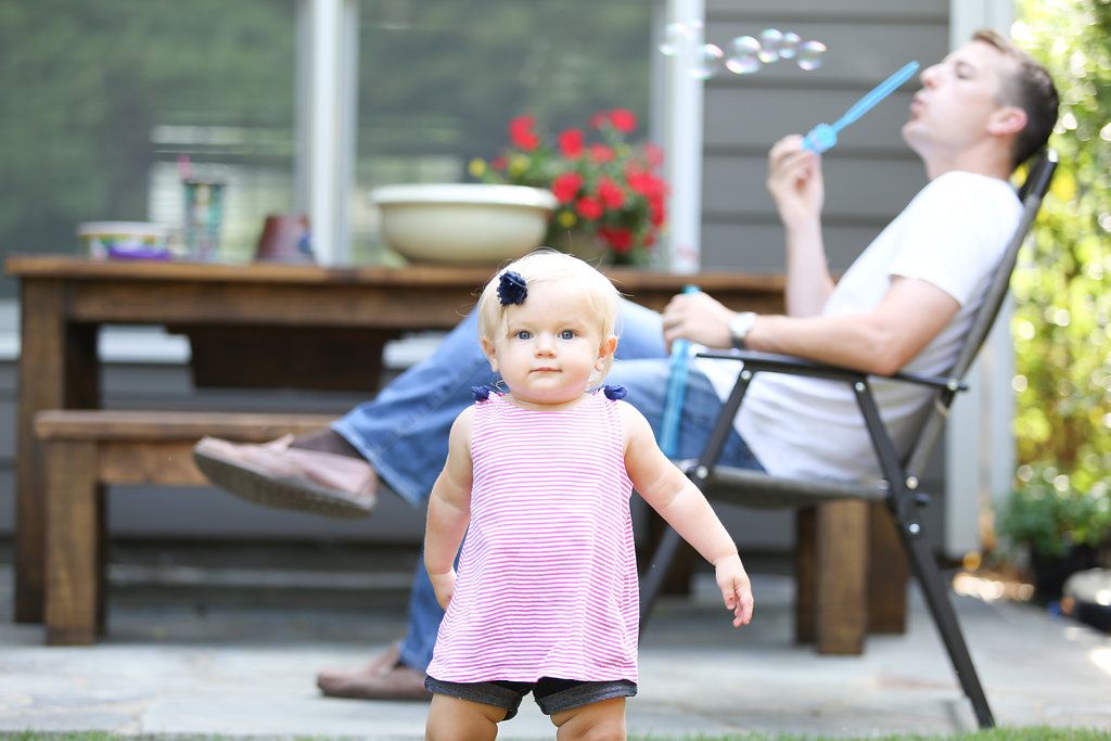 A toddler in a pink dress stands on a patio while a man relaxes in a chair blowing bubbles outdoors.