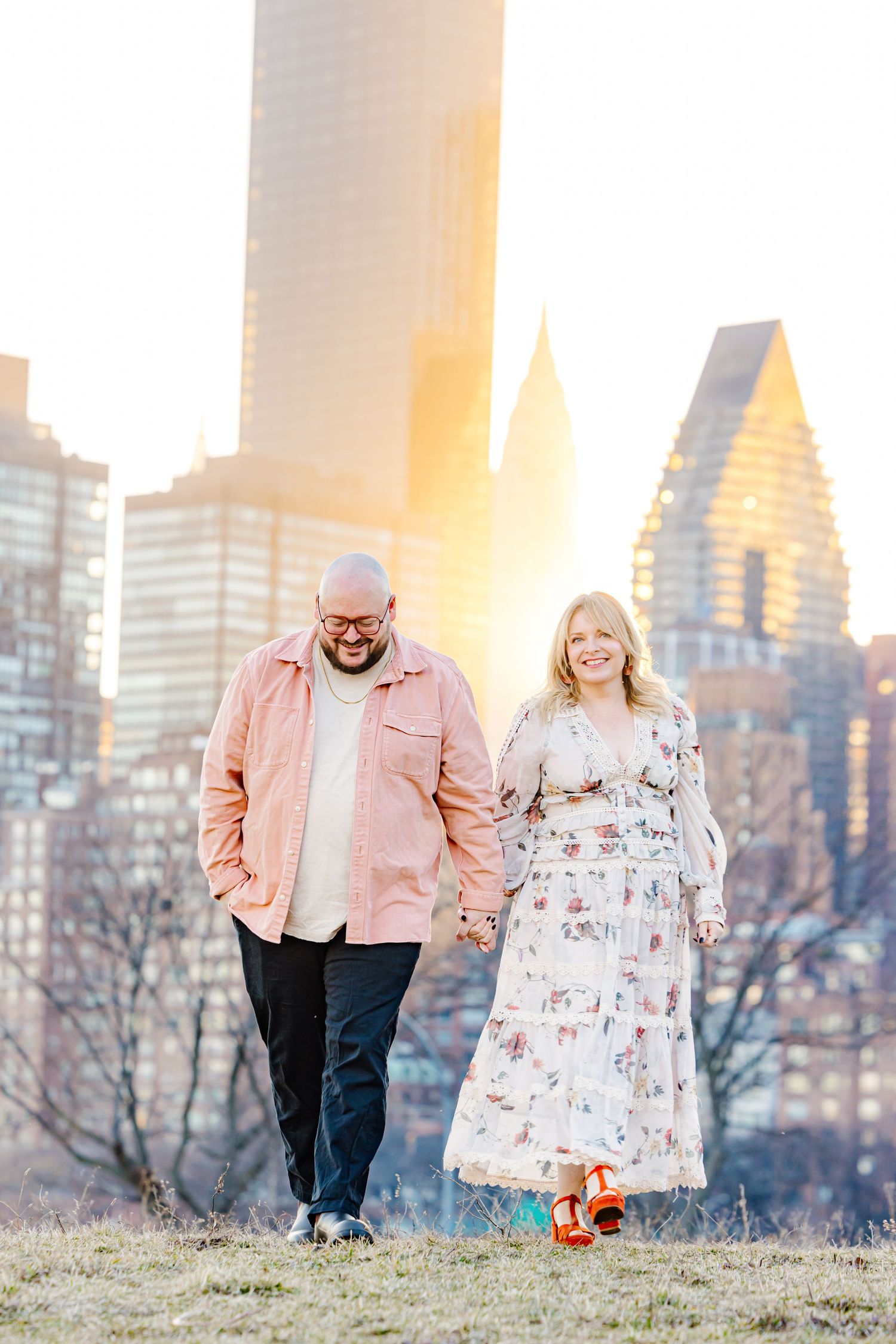 Couple holding hands and smiling while walking in a park with golden sunset light illuminating a city skyline behind them.