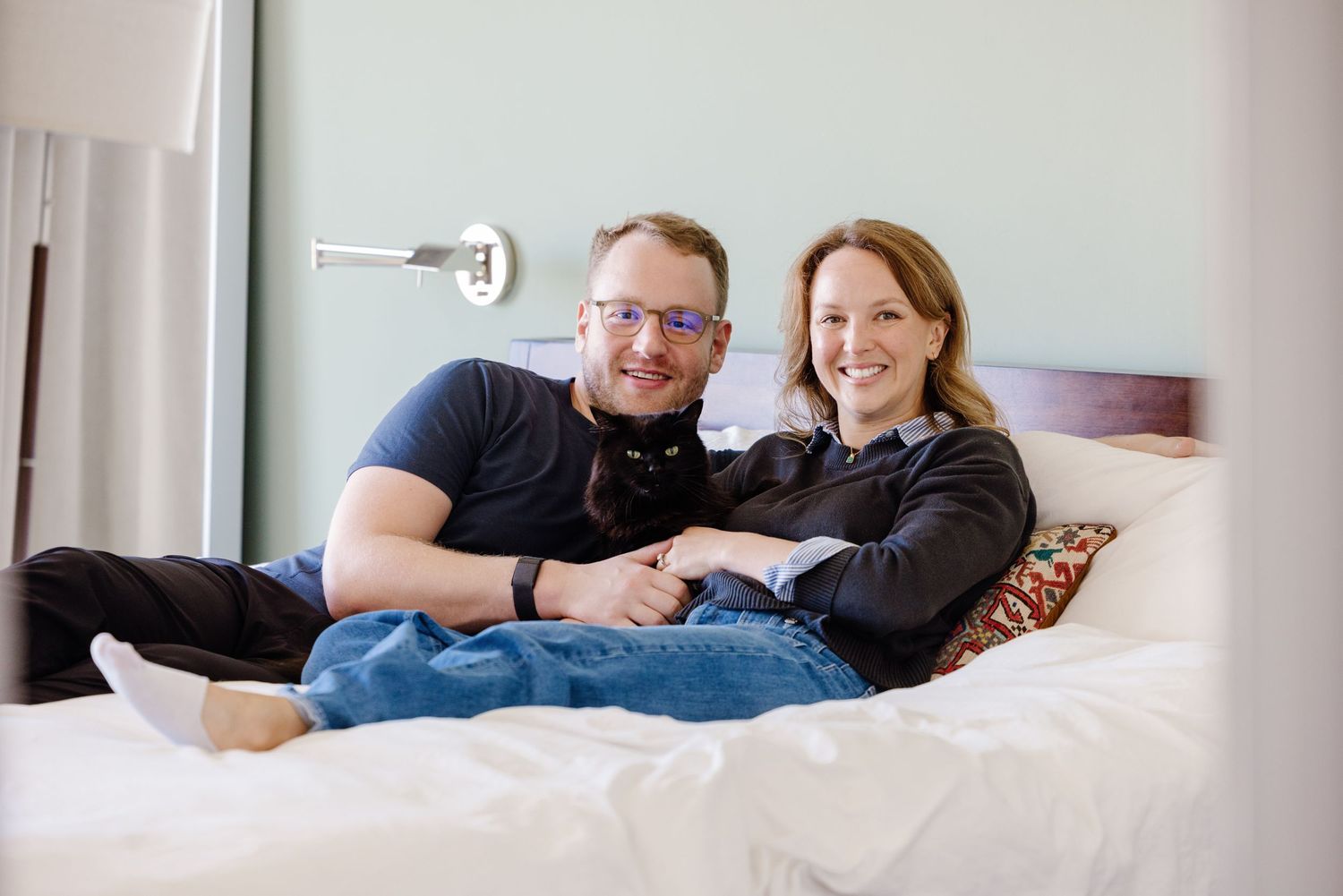 Couple relaxing on a white bed with their black cat, smiling warmly in a cozy bedroom setting.