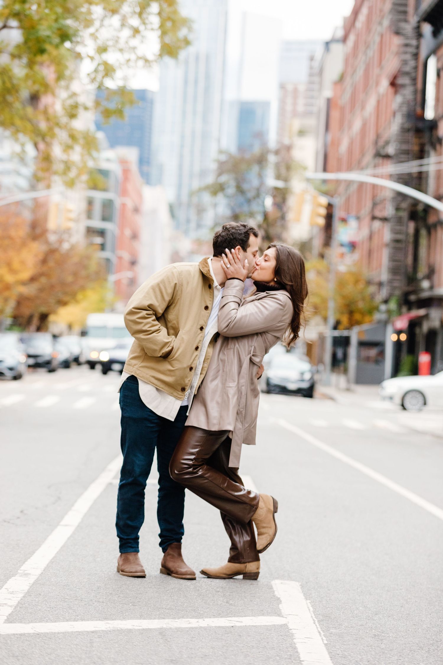 Couple sharing a passionate kiss on a city street surrounded by autumn trees and buildings.