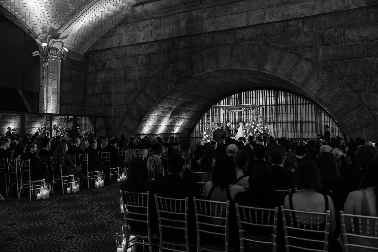 Black and white wide-angle shot of wedding ceremony under stone arches with guests seated in chiavari chairs.
