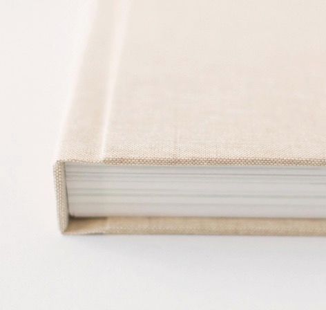 Close-up of a hardcover book with a beige linen fabric cover and thick white pages on a white surface.