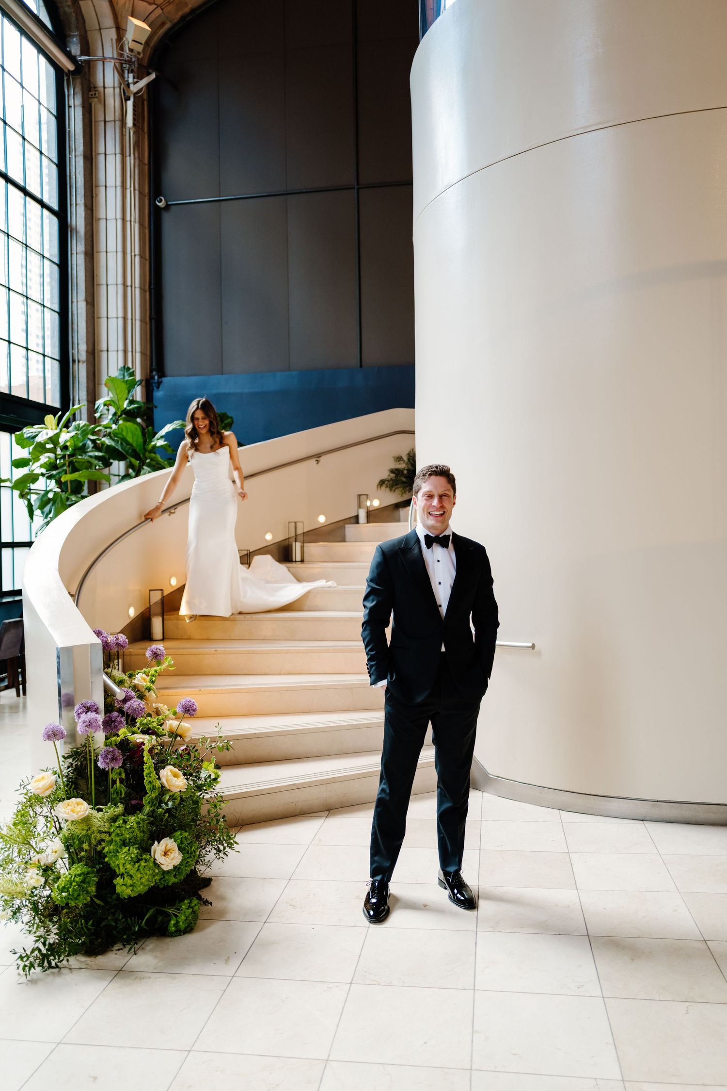 Bride descends curved staircase in white gown as groom in tuxedo stands smiling beside floral arrangement.