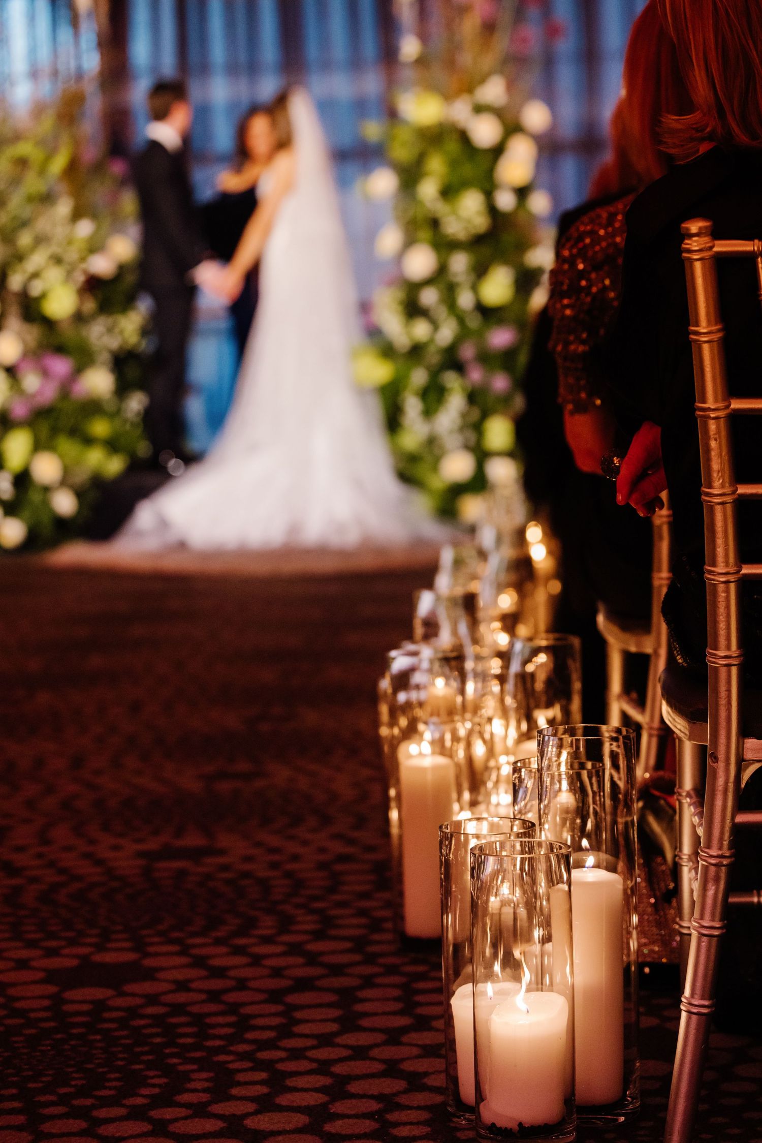 Candlelit wedding aisle with glass hurricane candle holders lining the path as couple embraces at the altar.