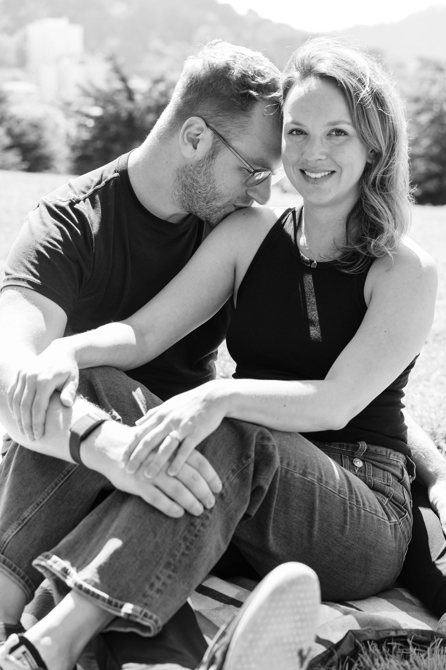 Black and white photo of a couple sitting outdoors, sharing a tender moment in the warm sunlight.
