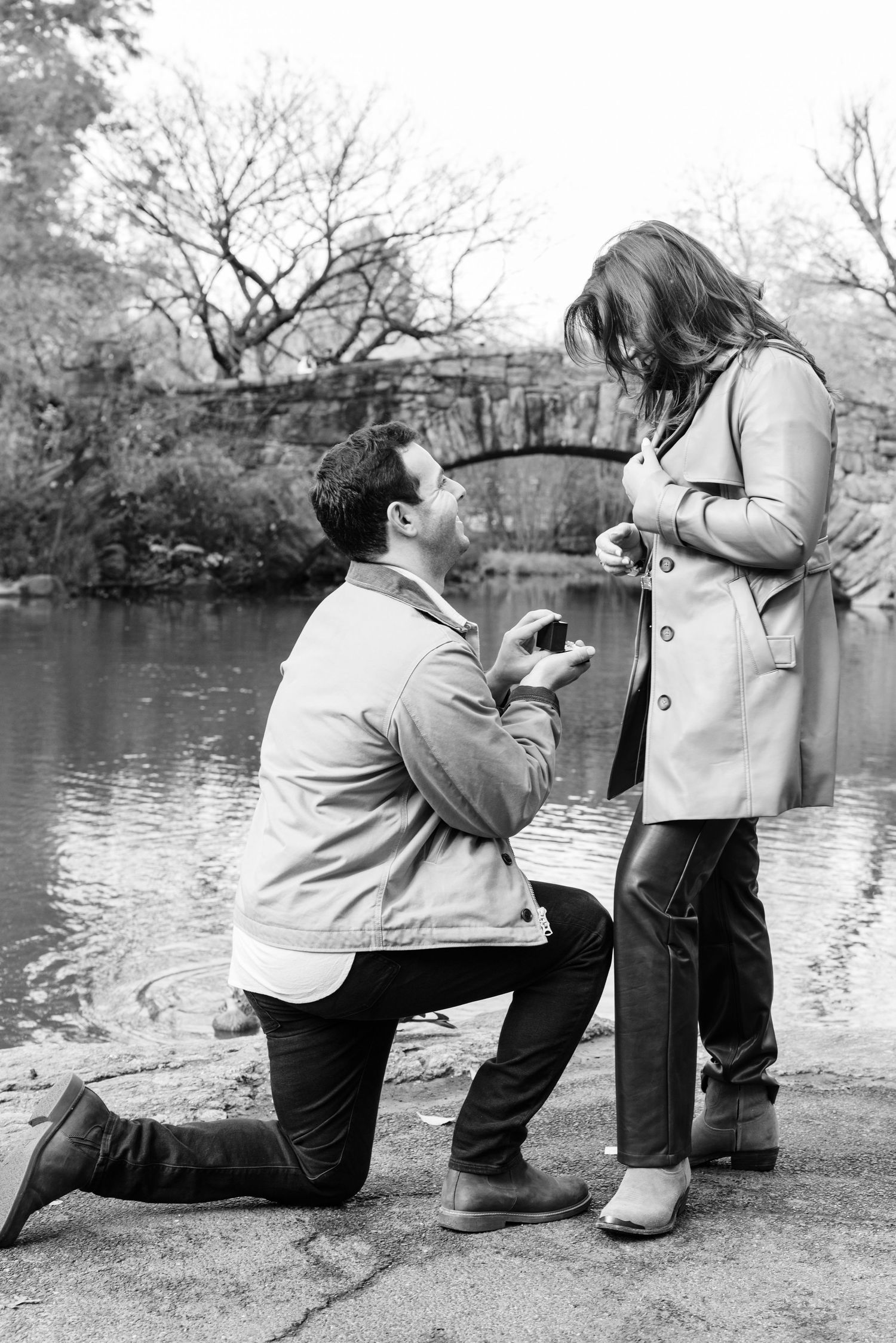 Black and white photo of man proposing on one knee to woman near a pond with stone bridge backdrop.