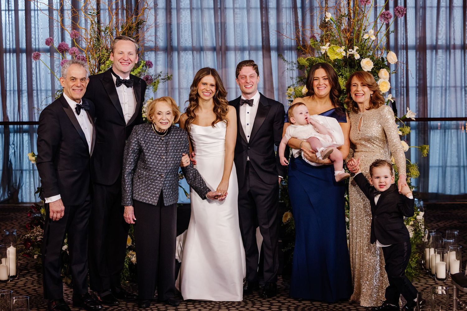 Wedding couple posing with extended family including baby and young child before floral altar.