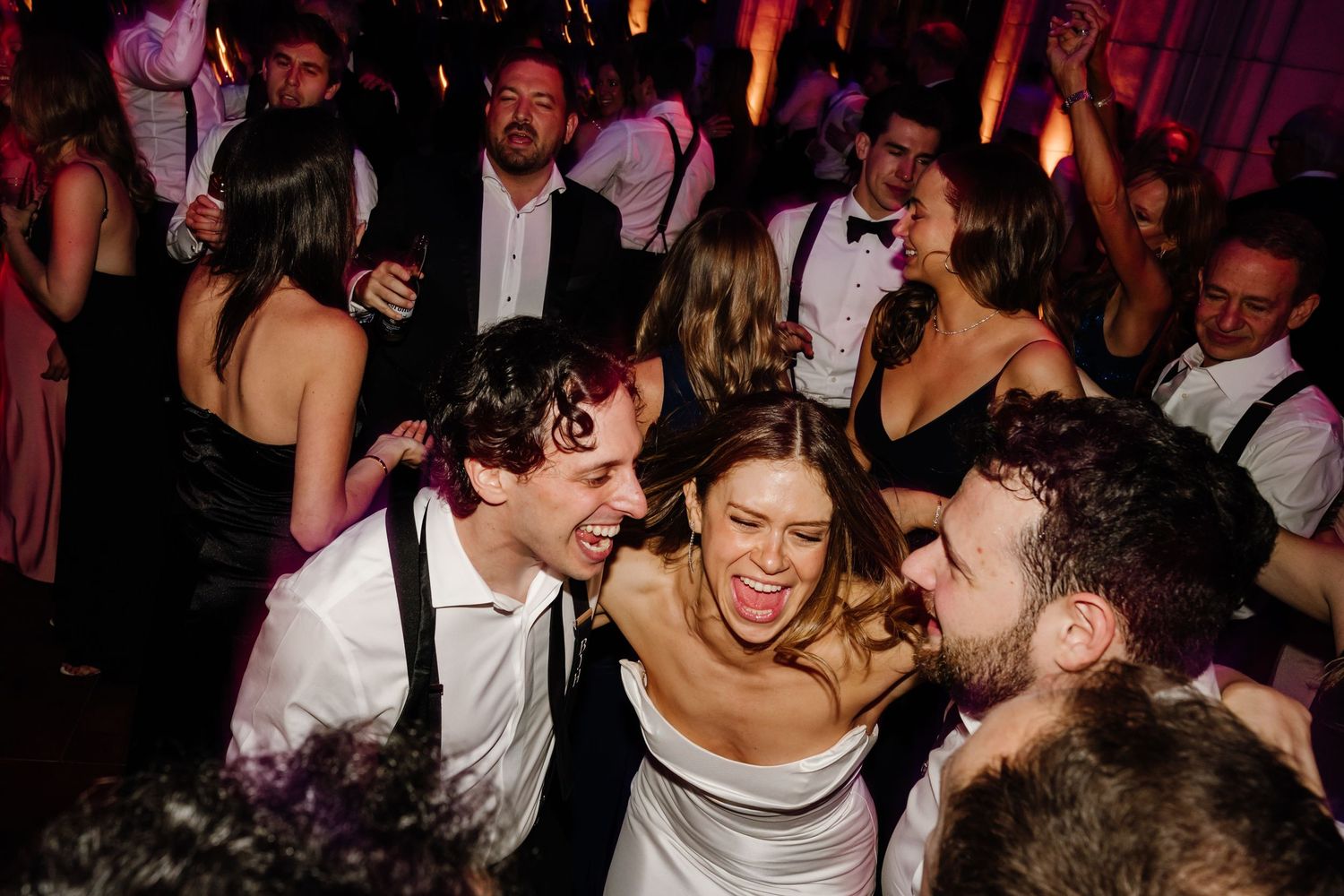 Bride and groom celebrating joyfully with friends on crowded wedding dance floor.
