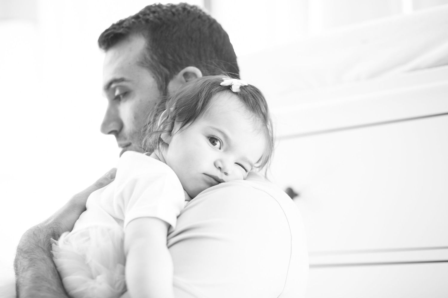 Black and white photo of a father tenderly holding his baby girl against his shoulder indoors.